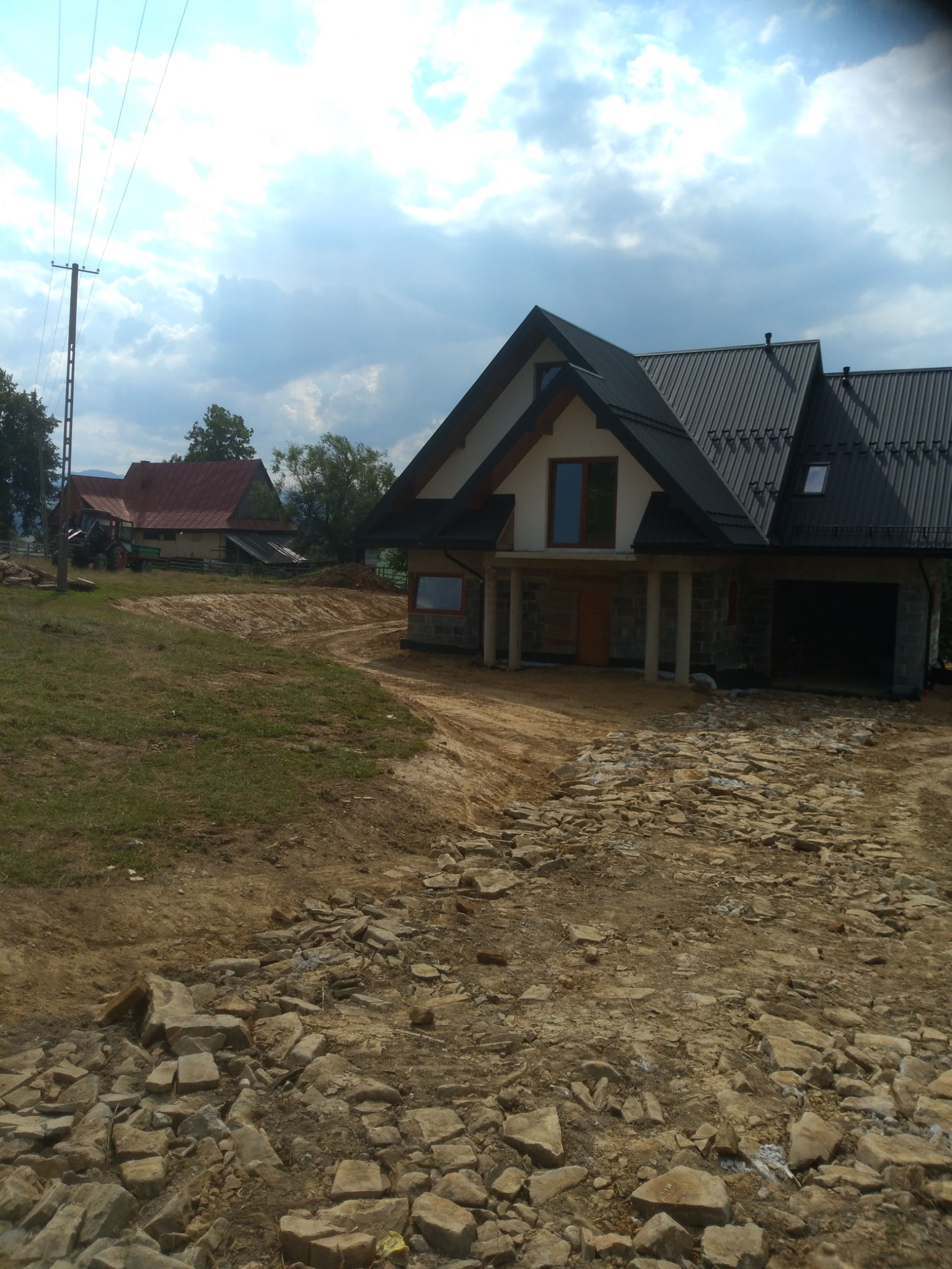 Nowo budowany dom z czarnym dachem i szarymi kamiennymi elementami, widoczny teren po robotach ziemnych i stary budynek w tle.