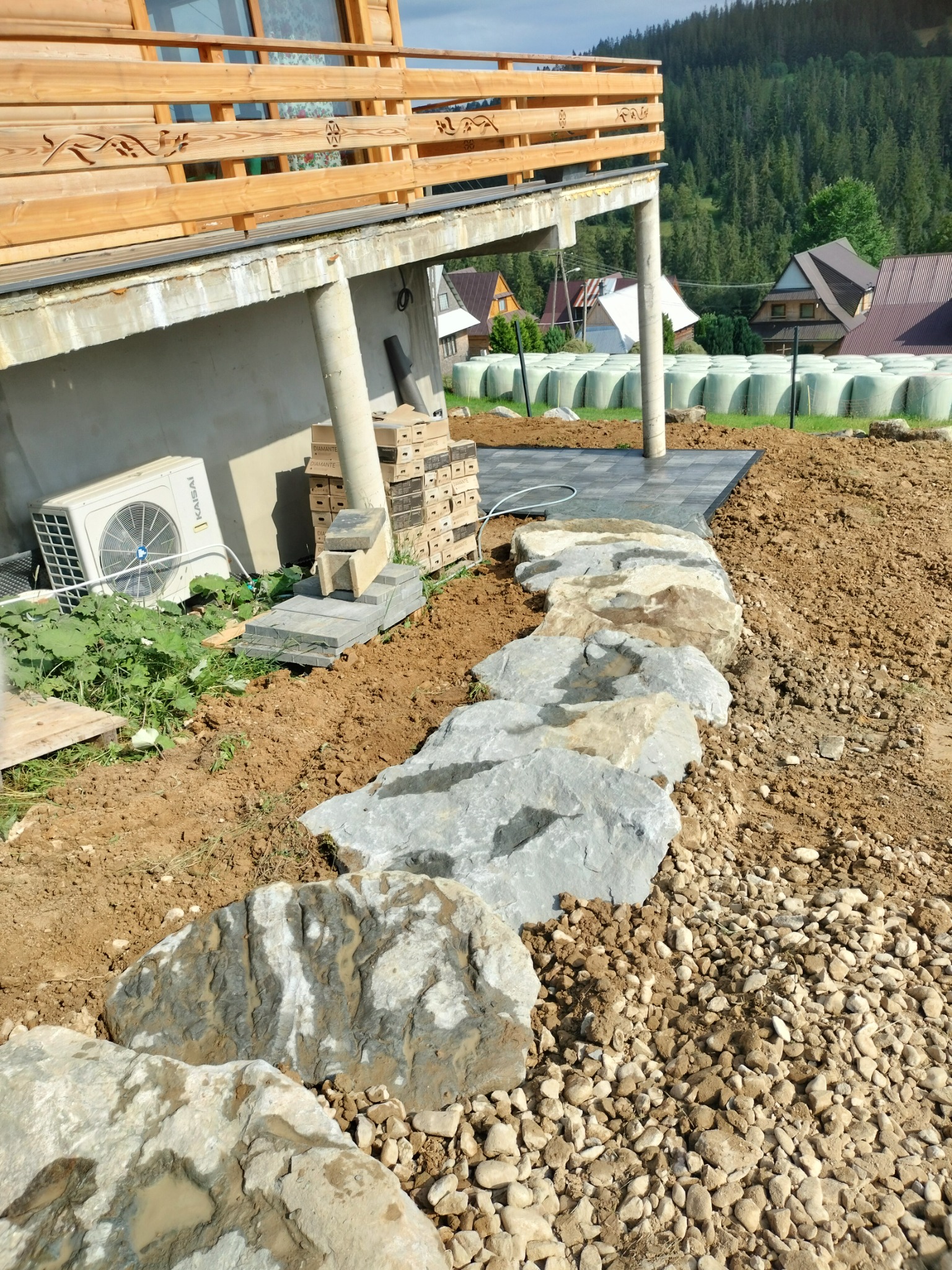 Układanie kamiennego murka oporowego z nieregularnych głazów na nierównym terenie, w tle widoczny dom z drewnianą balustradą i stogi siana.