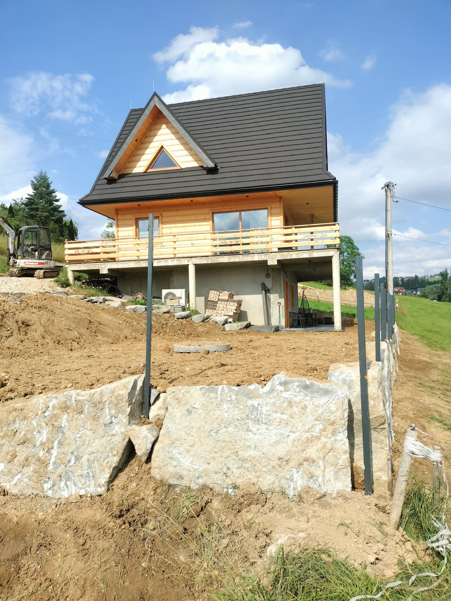 Nowoczesny dom z bali na wzniesieniu, widoczne prace ziemne, mur z kamienia i przygotowane słupki pod ogrodzenie, na drugim planie minikoparka.