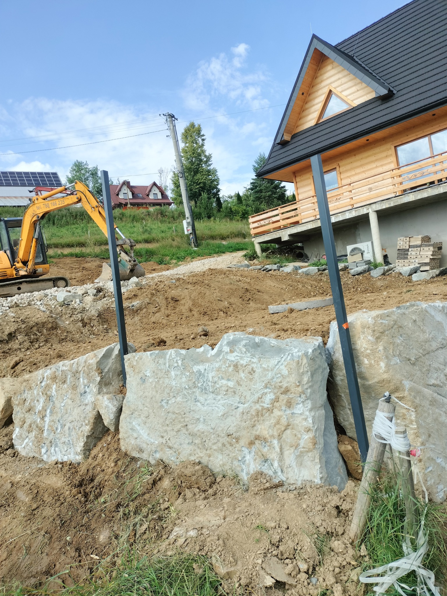 Żółta koparka przy budowie skalnego muru oporowego na nierównym terenie, z widokiem na drewniany dom z poddaszem i panele słoneczne w tle.