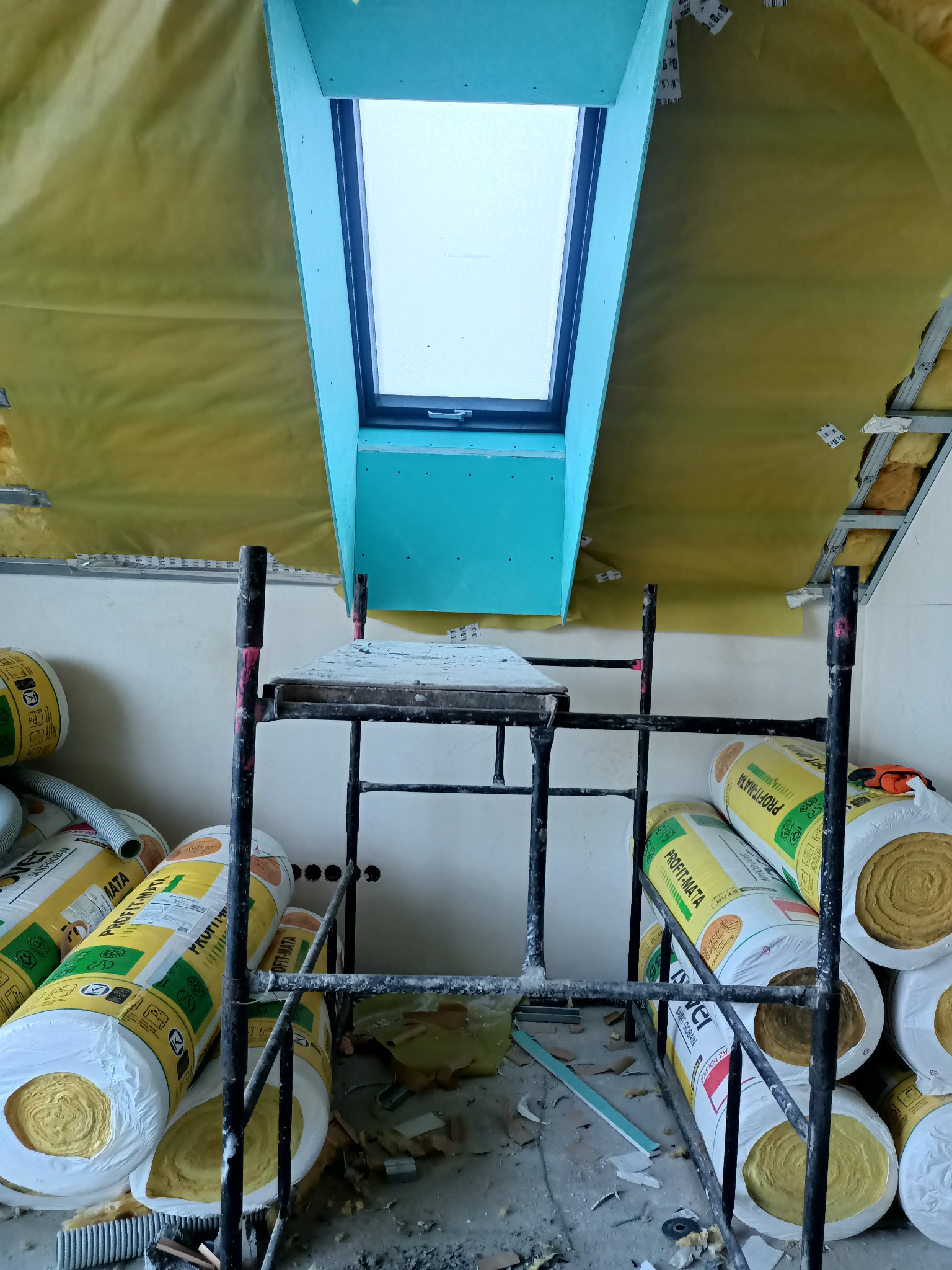 Poddasze w trakcie ocieplania z widocznym oknem dachowym w obudowie z płyt gipsowo-kartonowych, rusztowaniem i rolkami wełny mineralnej PROFIT-MATA.