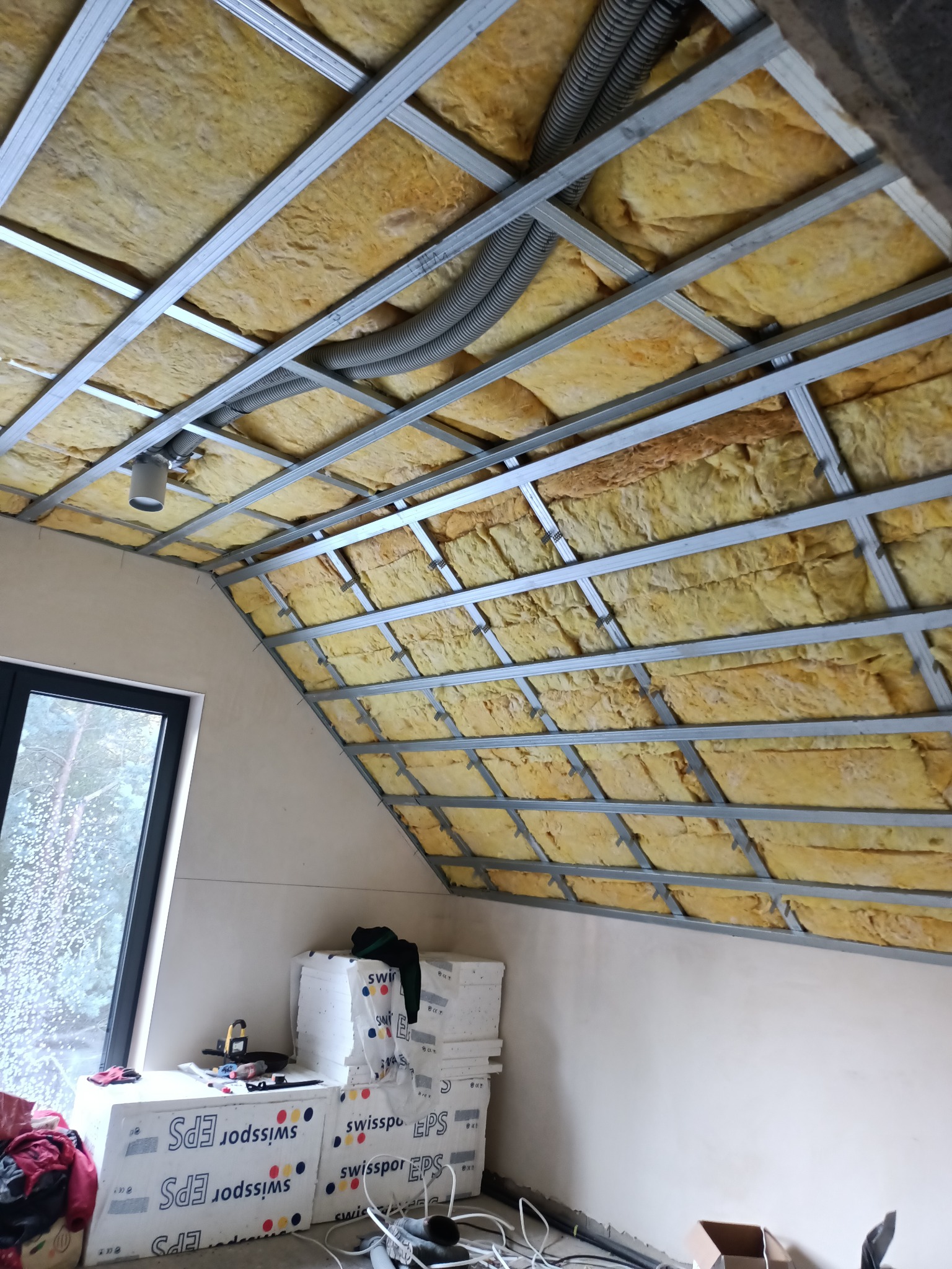 Poddasze w trakcie ocieplania wełną mineralną, widoczne metalowe profile sufitowe, wentylacja i okno dachowe.