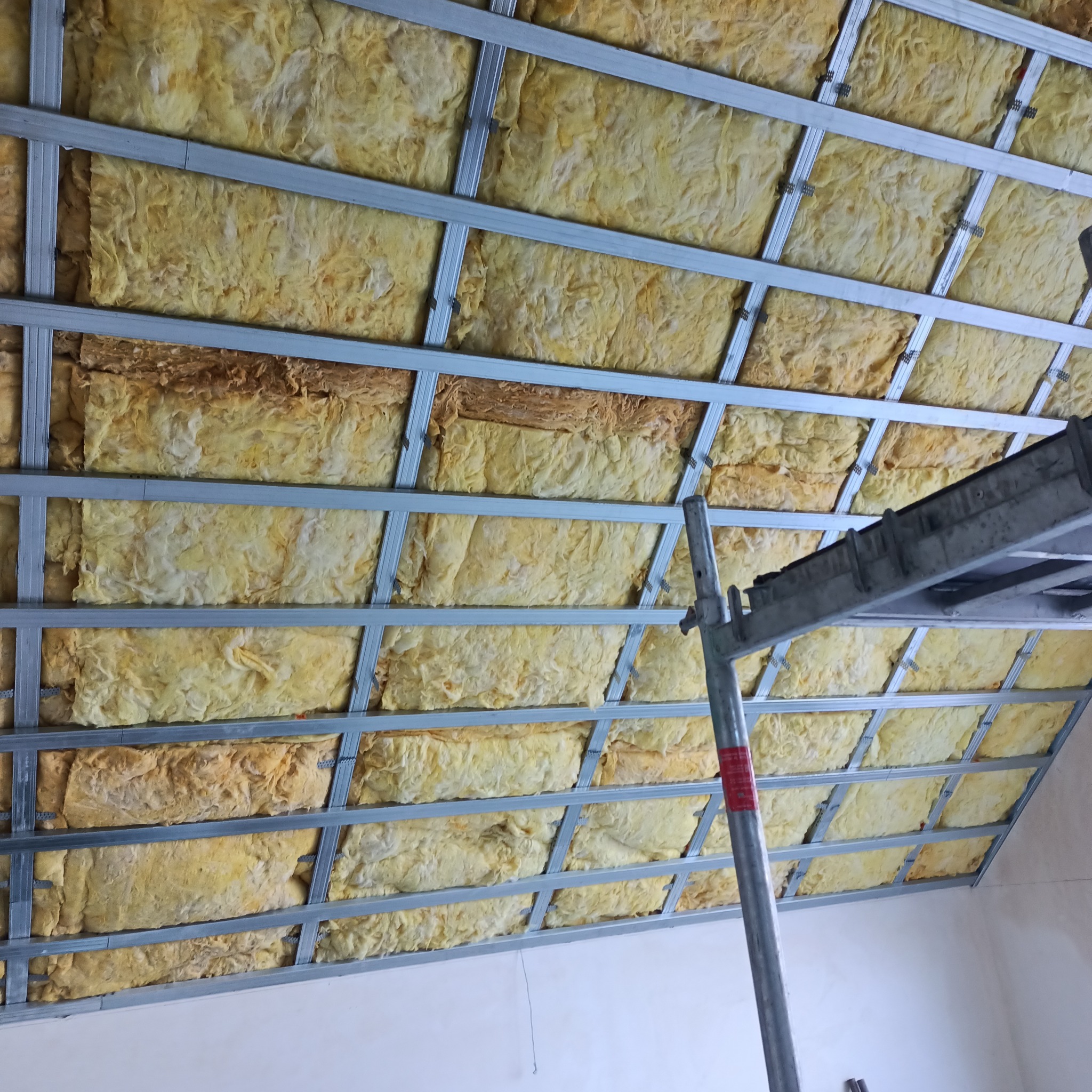 Żółta wełna mineralna w metalowym stelażu pod dachem, widok z dołu z fragmentem rusztowania.