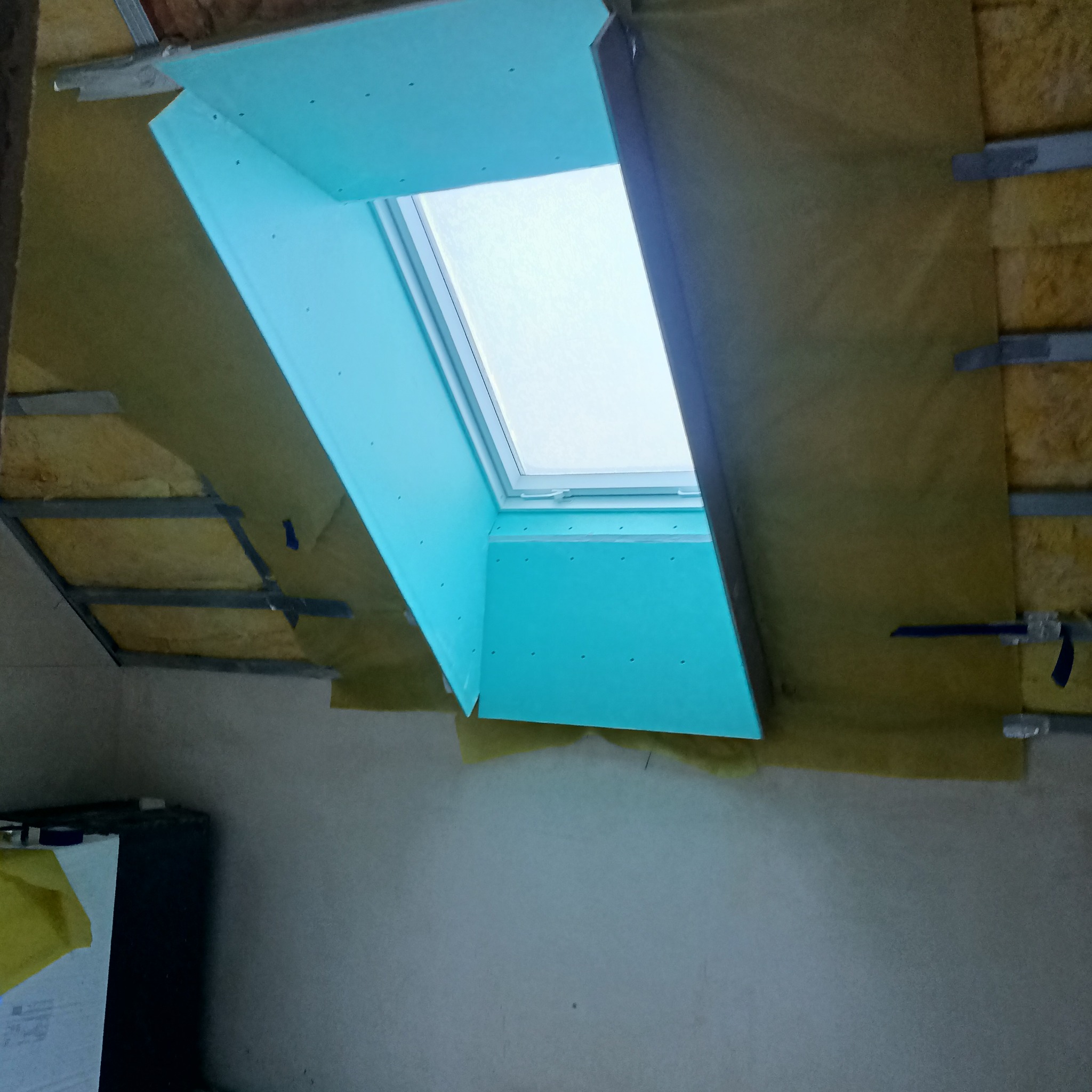 Montaż okna dachowego z obudową z płyty gipsowo-kartonowej w ocieplonym poddaszu, widoczne fragmenty wełny mineralnej i metalowego stelażu.