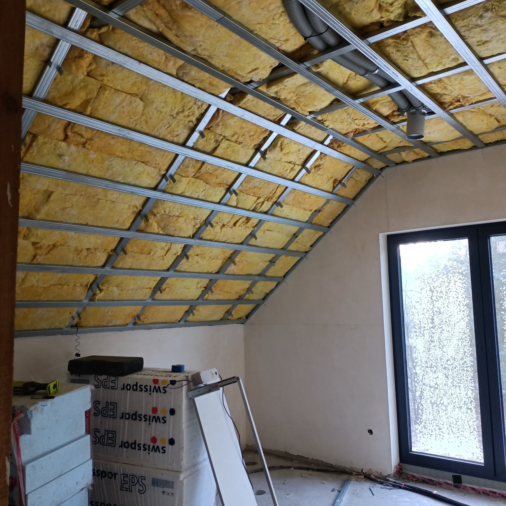 Skośne poddasze w trakcie ocieplania, widoczna wełna mineralna pomiędzy metalowymi profilami, w tle okno, na pierwszym planie paczki styropianu EPS.