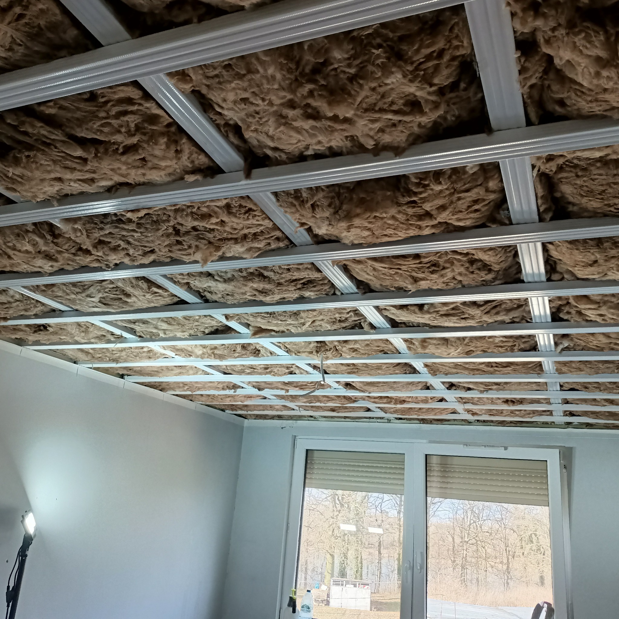 Izolacja z wełny mineralnej umieszczona w metalowym stelażu podwieszanego sufitu, widok z dołu, fragment okna w tle.