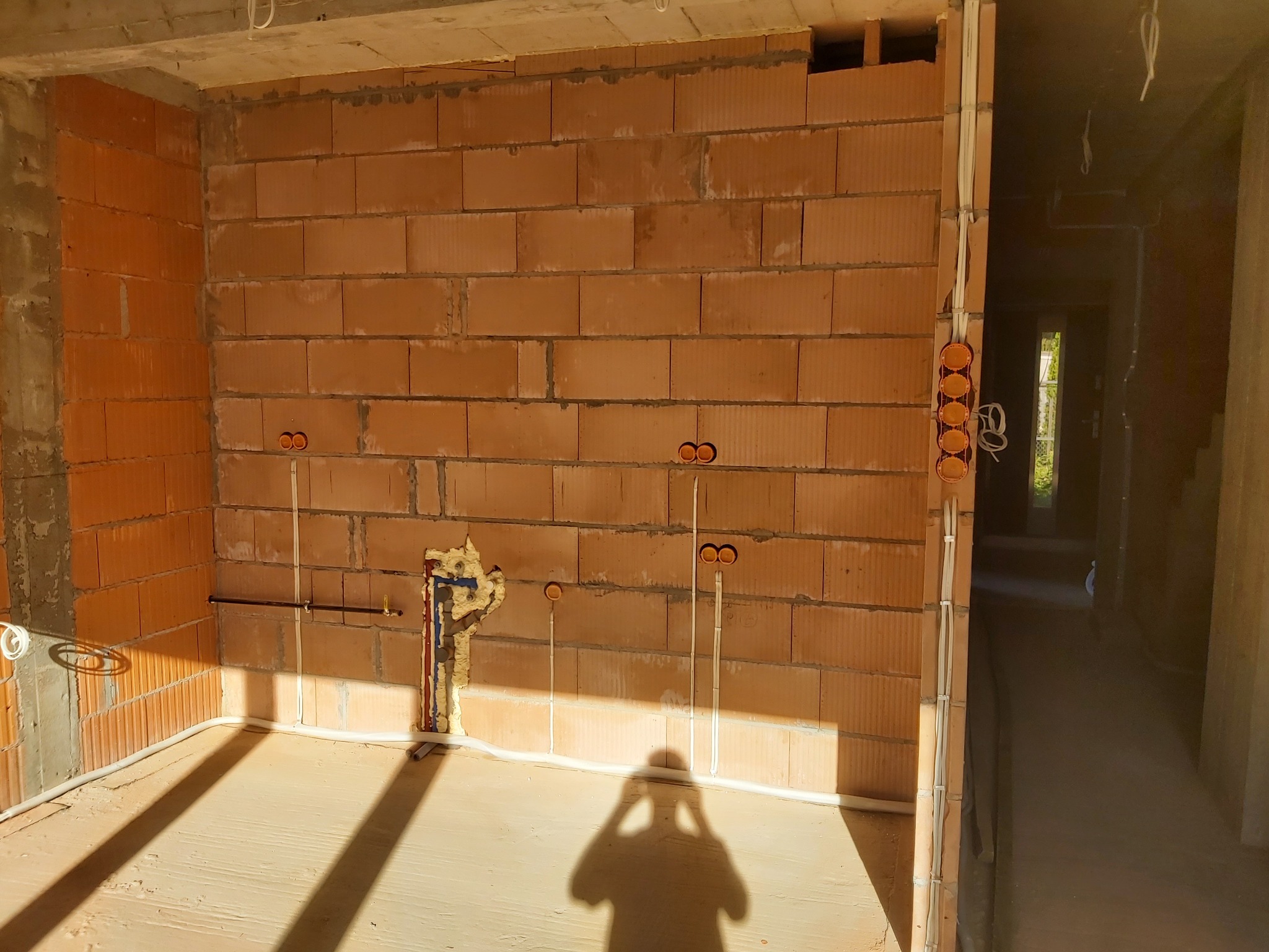 Surowy stan pomieszczenia z instalacją elektryczną w ścianach z cegły, widoczne puszki pod gniazdka, przewody oraz instalacja wodna zabezpieczona pianką montażową, cień fotografa na podłodze.