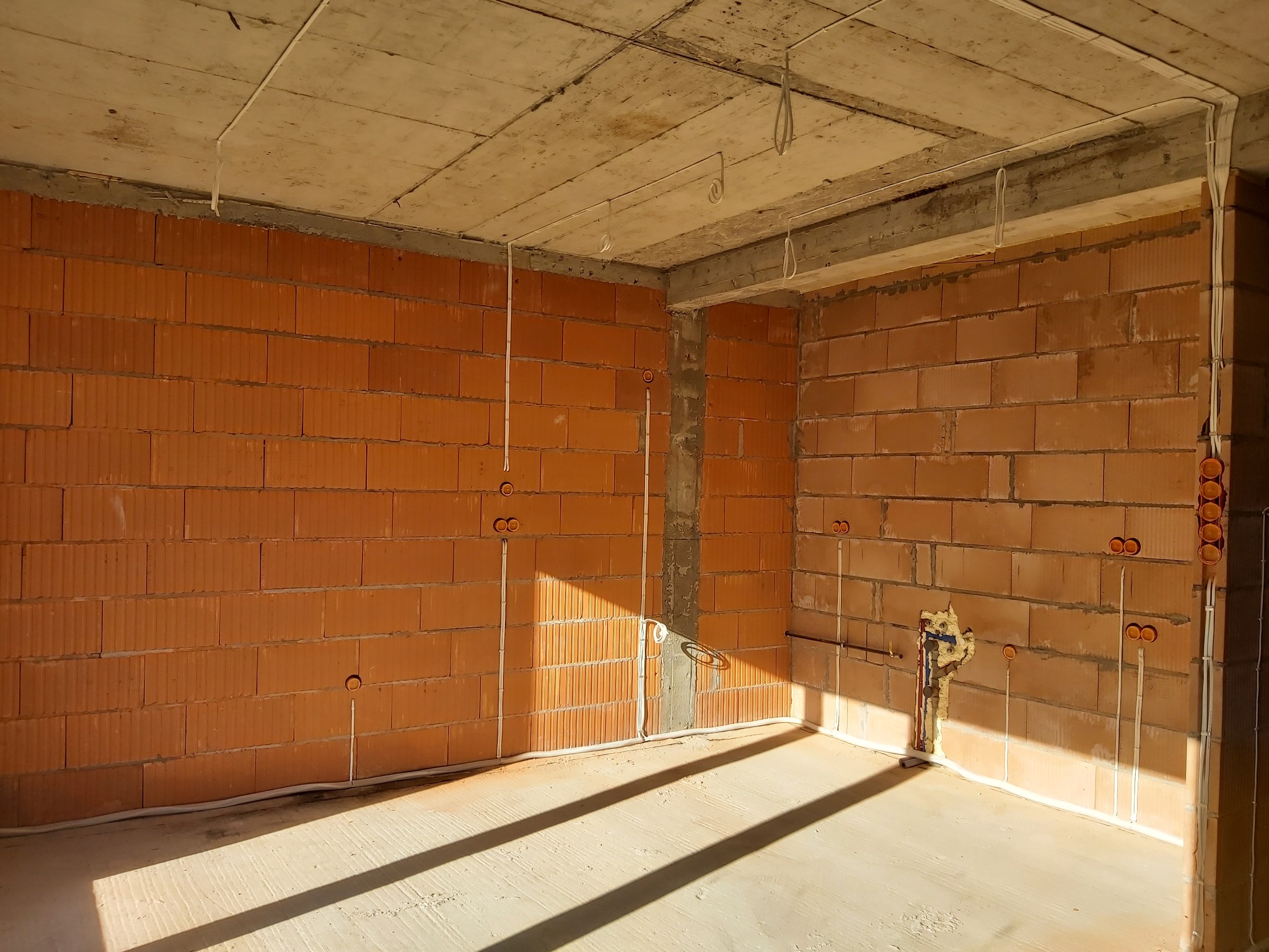 Surowy stan wnętrza z ceglanymi ścianami i poprowadzonymi przewodami elektrycznymi, widoczne puszki instalacyjne w ścianach oraz przewody pod sufitem, w tle ściana z instalacją wodną zabezpieczoną...