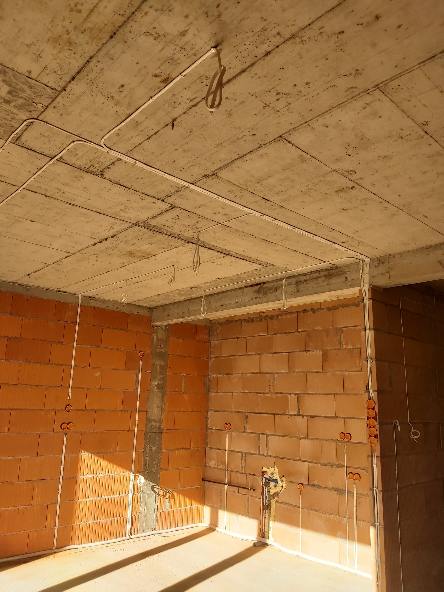Surowy stan wnętrza z ceglanymi ścianami, betonowym stropem i rozprowadzonymi przewodami elektrycznymi w białych rurkach PCV, widoczne puszki instalacyjne w kolorze pomarańczowym.