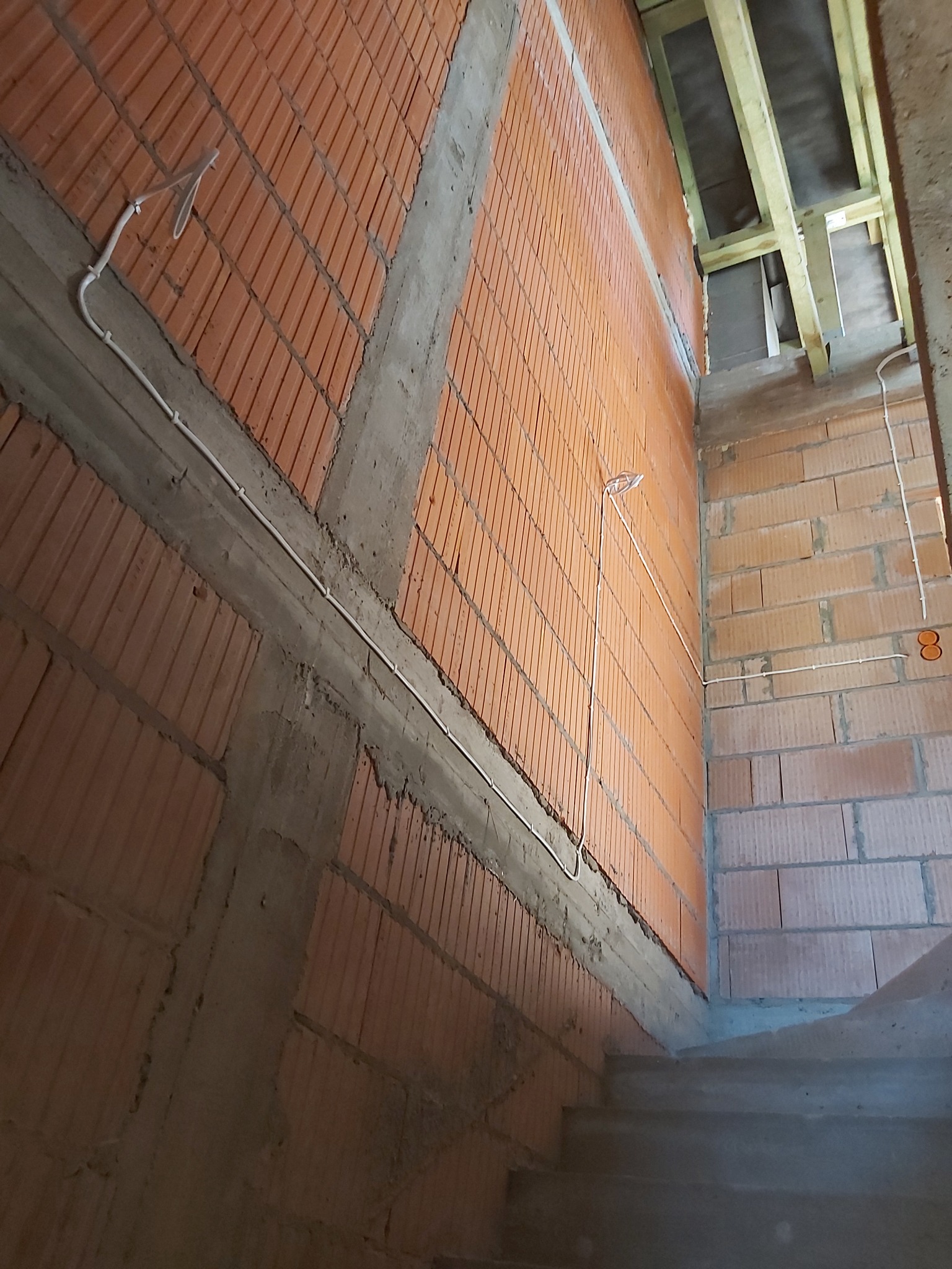 Surowe wnętrze w trakcie budowy: widoczne ceglane ściany, betonowe wzmocnienia, schody i prowadzone przewody elektryczne w białych rurkach.