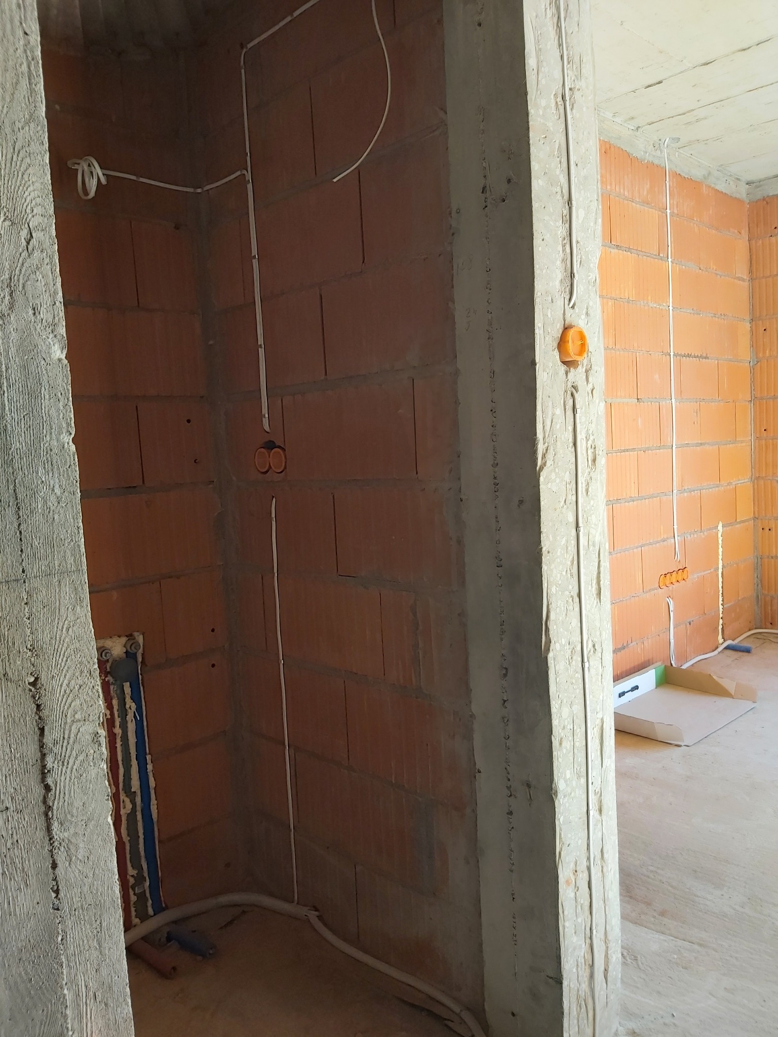 Surowy stan: ściany z czerwonej cegły dziurawki z widocznym pionowym prowadzeniem przewodów elektrycznych w białych rurkach, puszki instalacyjne w ścianie, fragment pionu wodnego z rurami...