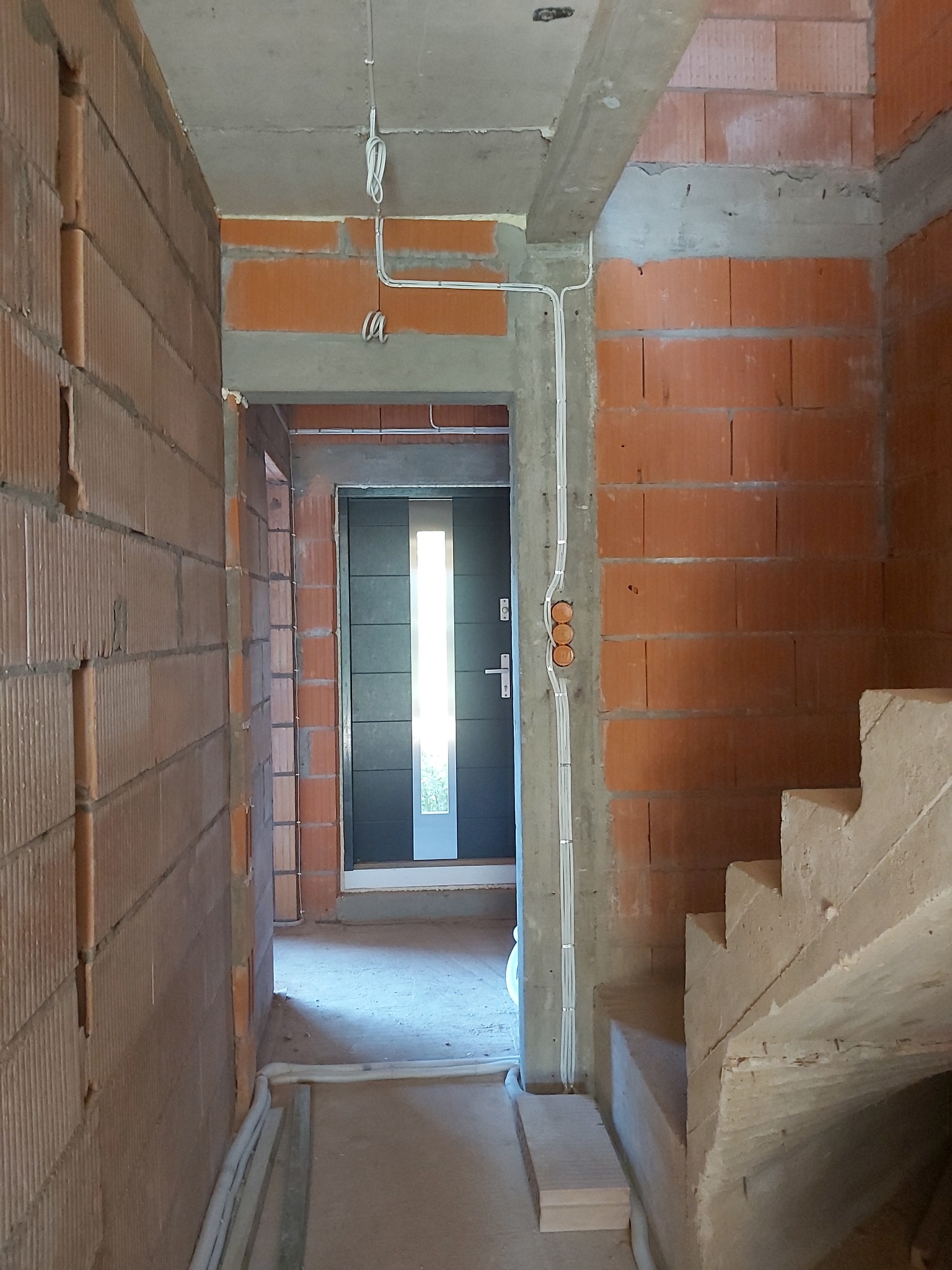 Wnętrze budynku w trakcie budowy: widoczne ściany z czerwonej cegły, betonowe schody, drzwi wejściowe z szybą i instalacja elektryczna w białych rurkach przytwierdzona do ściany.