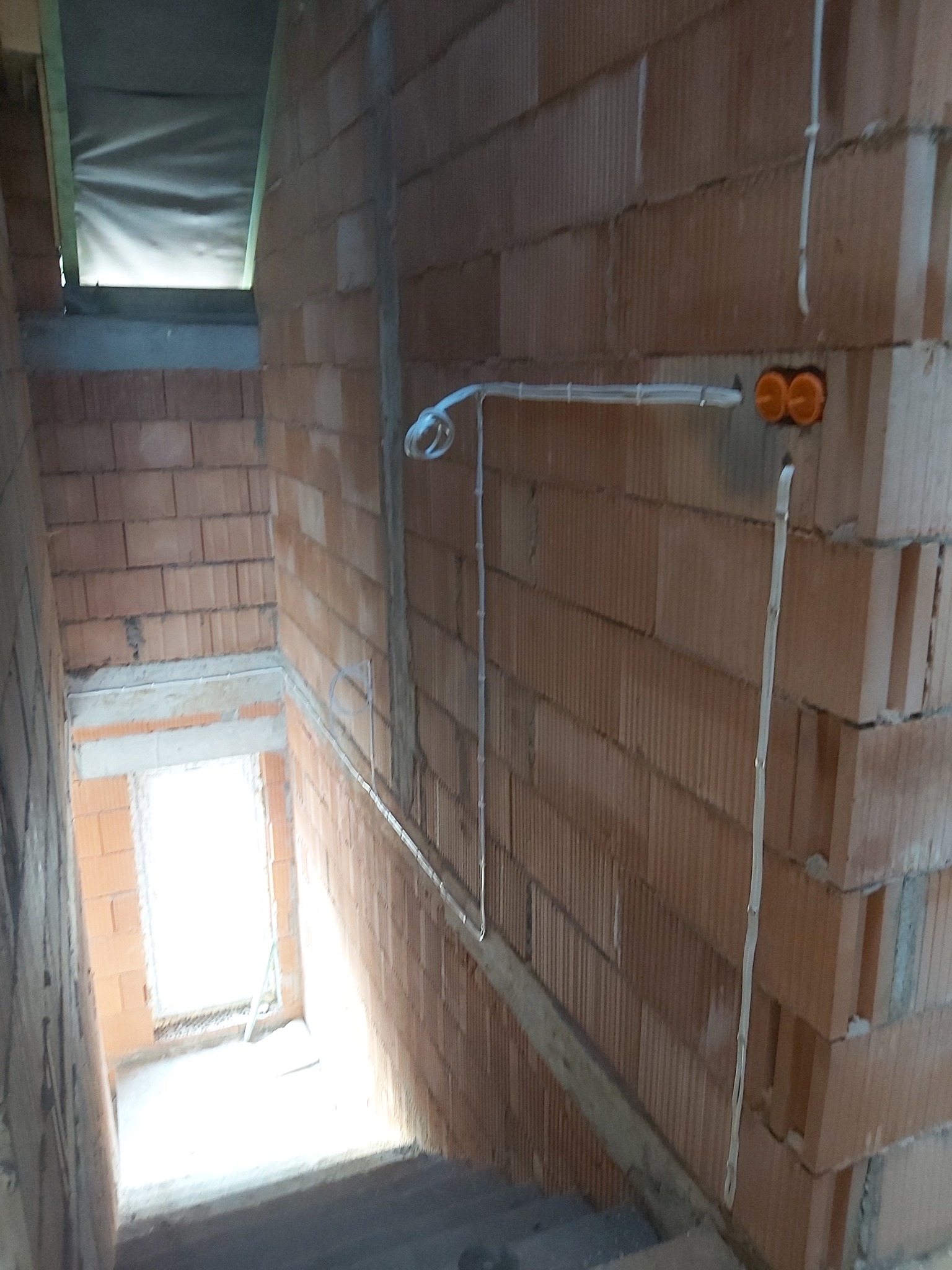 Kable elektryczne prowadzone po ścianie z cegły dziurawki w surowym stanie, widoczne puszki instalacyjne koloru pomarańczowego, w tle schody i okno z silnym światłem.