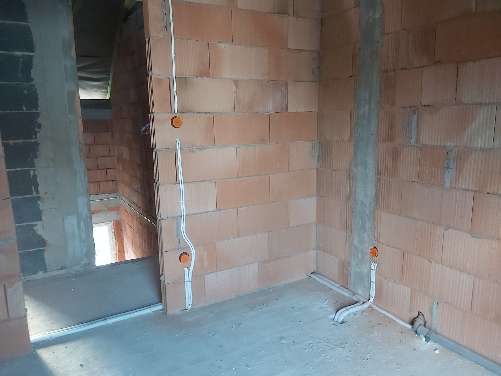 Wewnętrzne ściany z cegły dziurawki w surowym stanie z poprowadzonymi przewodami elektrycznymi w białych rurkach i pomarańczowymi puszkami instalacyjnymi, widok na sąsiednie pomieszczenie.
