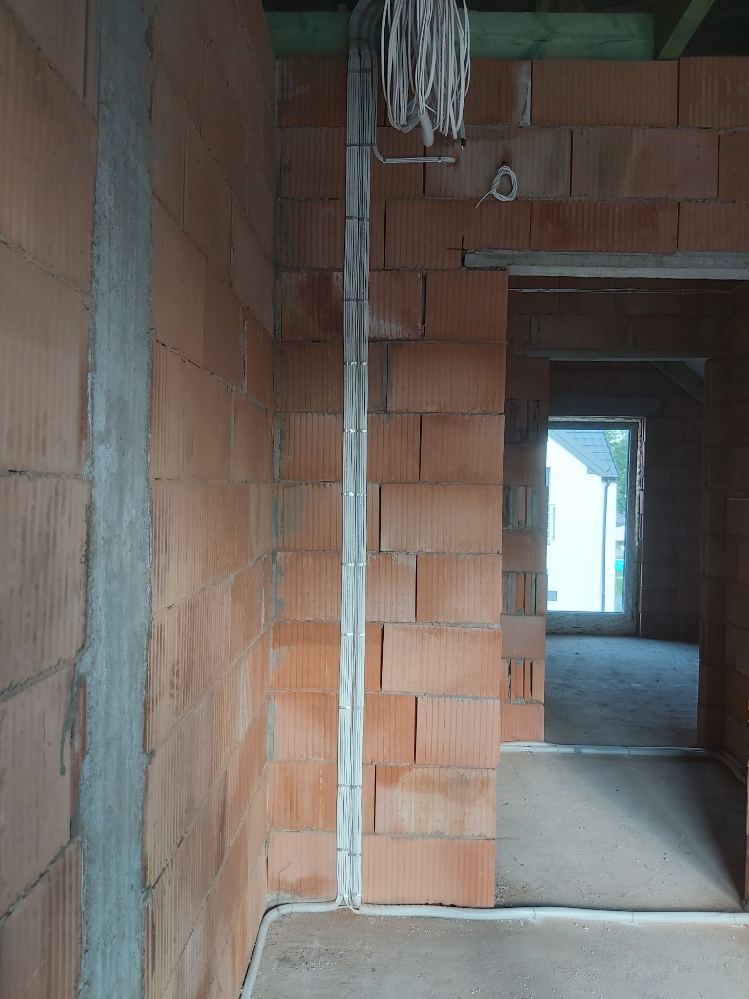 Surowe mury z czerwonej cegły ceramicznej, pionowy ciąg przewodów elektrycznych w białych listwach przymocowanych do ściany, kłębek kabli zwisający z góry, widok na sąsiednie pomieszczenie przez...