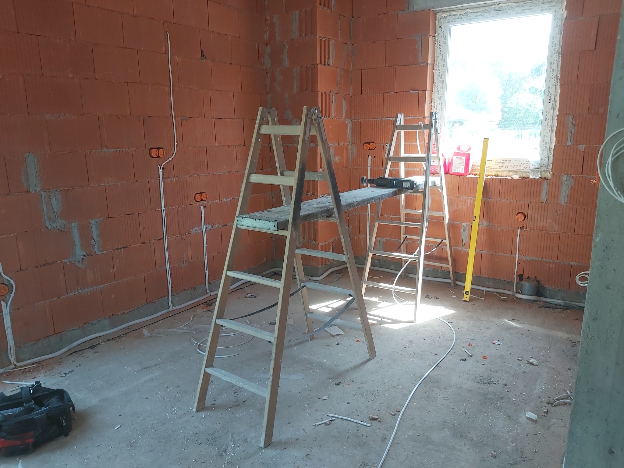 Wnętrze w stanie surowym z widocznymi przewodami elektrycznymi w ścianach z cegły, drabiny z podestem, poziomica oraz torba narzędziowa na podłodze.