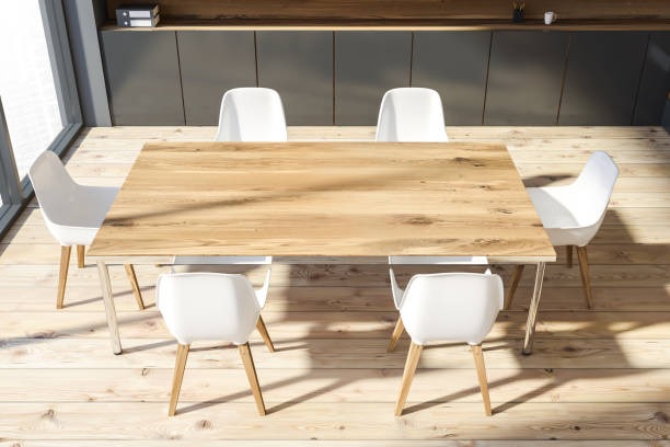 Blat drewniany stołu jadalnego z widocznym usłojeniem, otoczony białymi krzesłami na drewnianej podłodze, z szarą zabudową w tle.