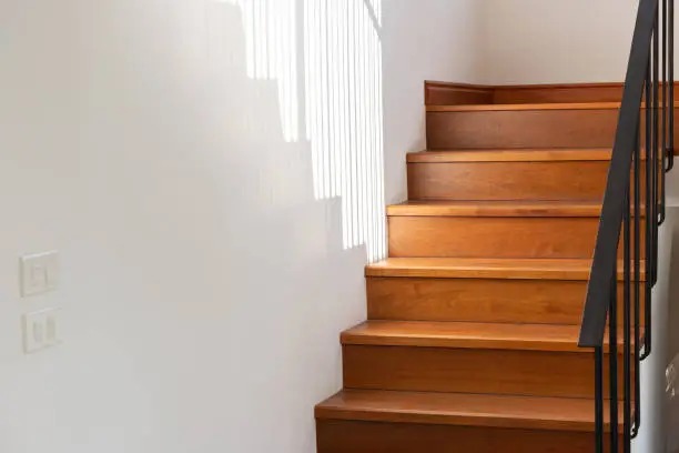 Drewniane schody z czarną metalową balustradą, oświetlone promieniami słonecznymi padającymi na białą ścianę obok.