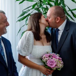 Martyna Baster - Photo - Panna młoda w białej sukience z bukietem całuje pana młodego w garniturze, przygląda im się uśmiechnięty mężczyzna w niebieskiej marynarce i wzorzystym krawacie, w tle roślina doniczkowa.