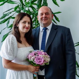 Martyna Baster - Photo - Para młoda pozuje do zdjęcia w pomieszczeniu z rośliną w tle. Panna młoda w białej sukience z bukietem różowych kwiatów, pan młody w granatowym garniturze i niebieskim krawacie.