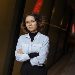 Martyna Baster - Photo - Portret młodej kobiety w okularach, ubrana w białą koszulę i czarny golf, pozująca na tle nowoczesnego budynku z ciemnymi panelami.