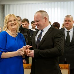 Martyna Baster - Photo - Uśmiechnięta para wymienia się obrączkami podczas ceremonii ślubnej w obecności gości, kobieta w niebieskiej sukience, mężczyzna w czarnym garniturze.