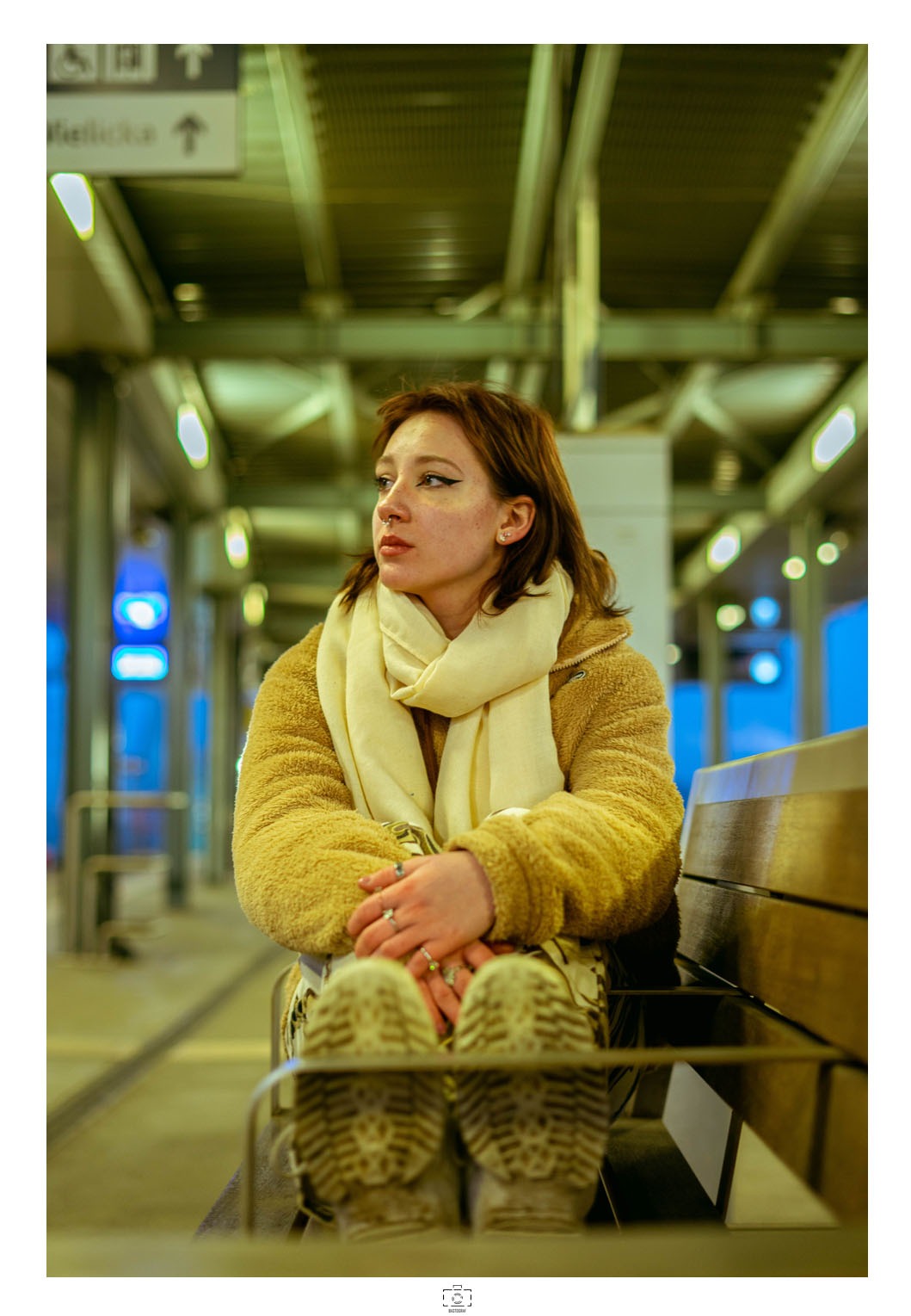 Portret młodej kobiety siedzącej na ławce na stacji, ubrana w futrzaną kurtkę i kremowy szal, z widocznymi butami i pierścionkami na dłoniach.