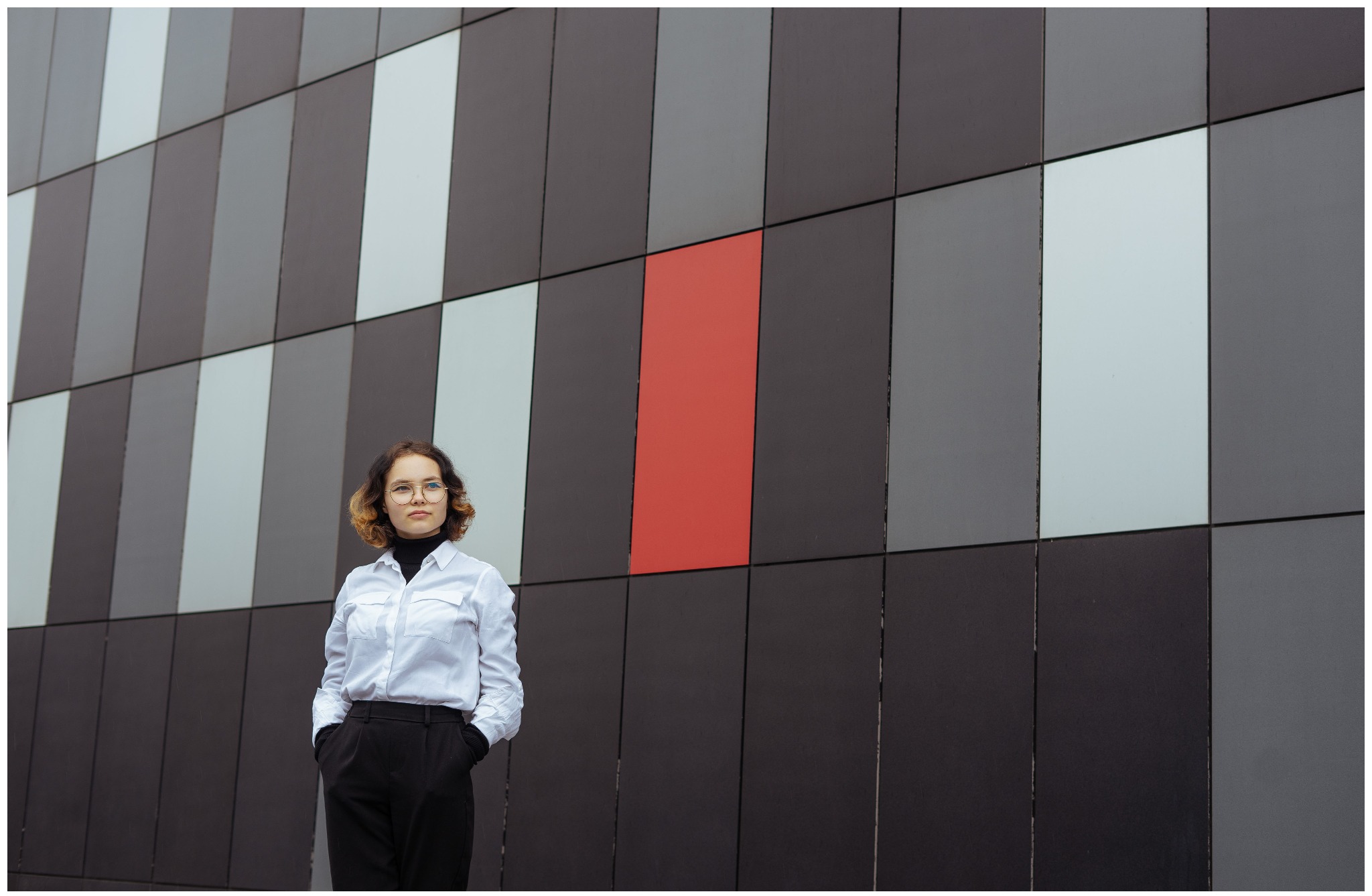 Portret młodej kobiety w okularach, stojącej na tle nowoczesnej fasady budynku z panelami w odcieniach szarości i czerwieni, ręce w kieszeniach spodni.