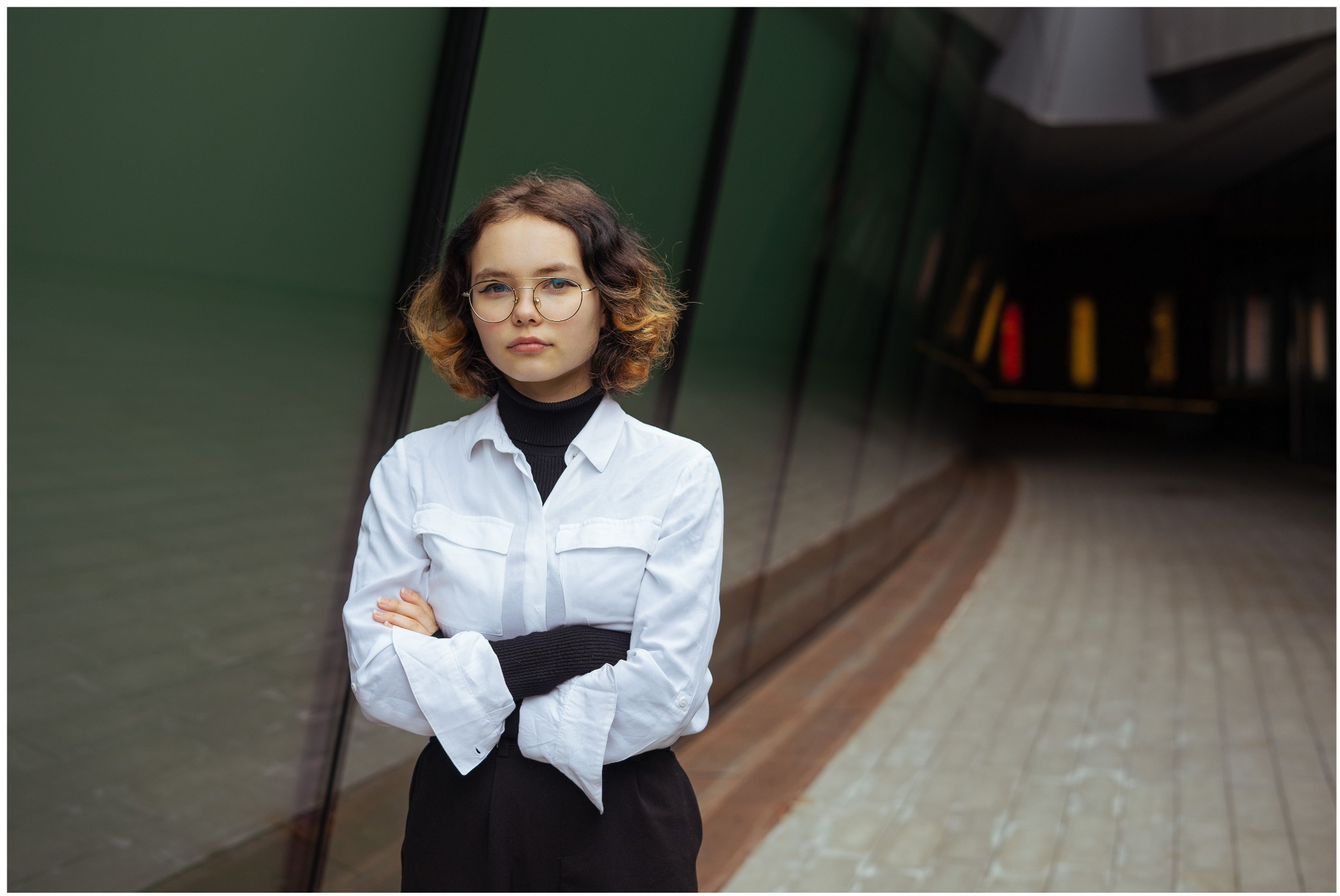 Młoda kobieta w okularach, czarnym golfie i białej koszuli stojąca na tle zielonej ściany i tunelu.