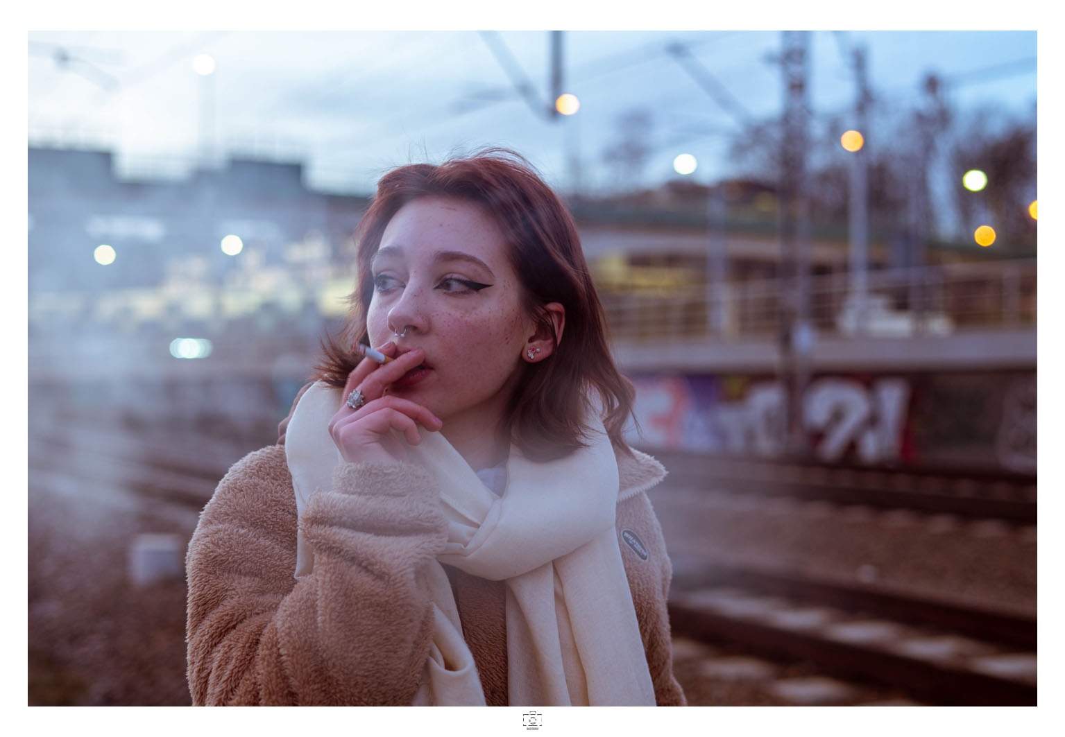 Portret młodej kobiety z papierosem na tle torów kolejowych w Krakowie, ubrana w beżową kurtkę i biały szal, z widocznymi kolczykami i pierścionkiem.