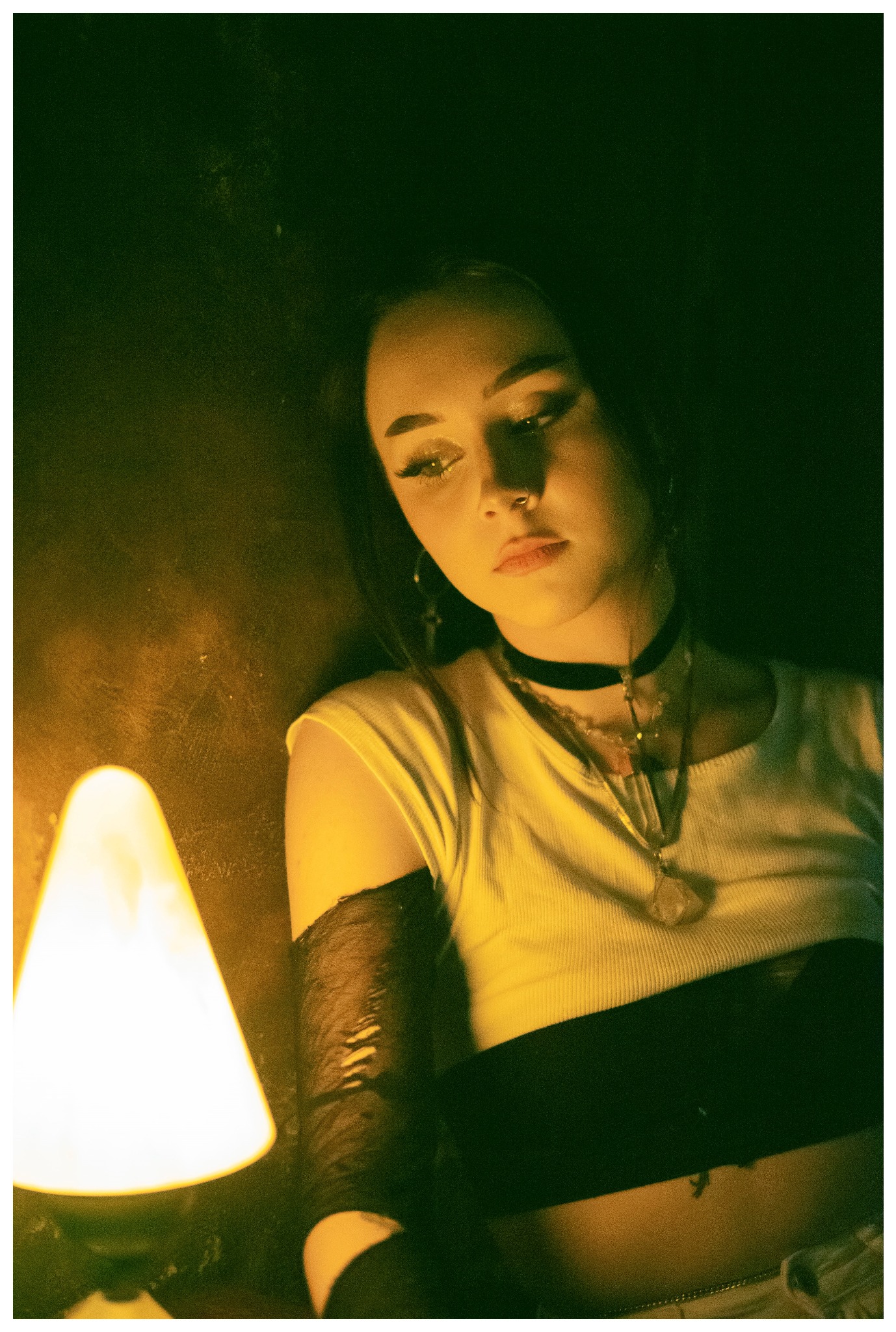 Portret młodej kobiety w klimatycznym, żółtym oświetleniu, z chokerem i naszyjnikami, zamyślona poza, widoczna lampka nocna w tle.