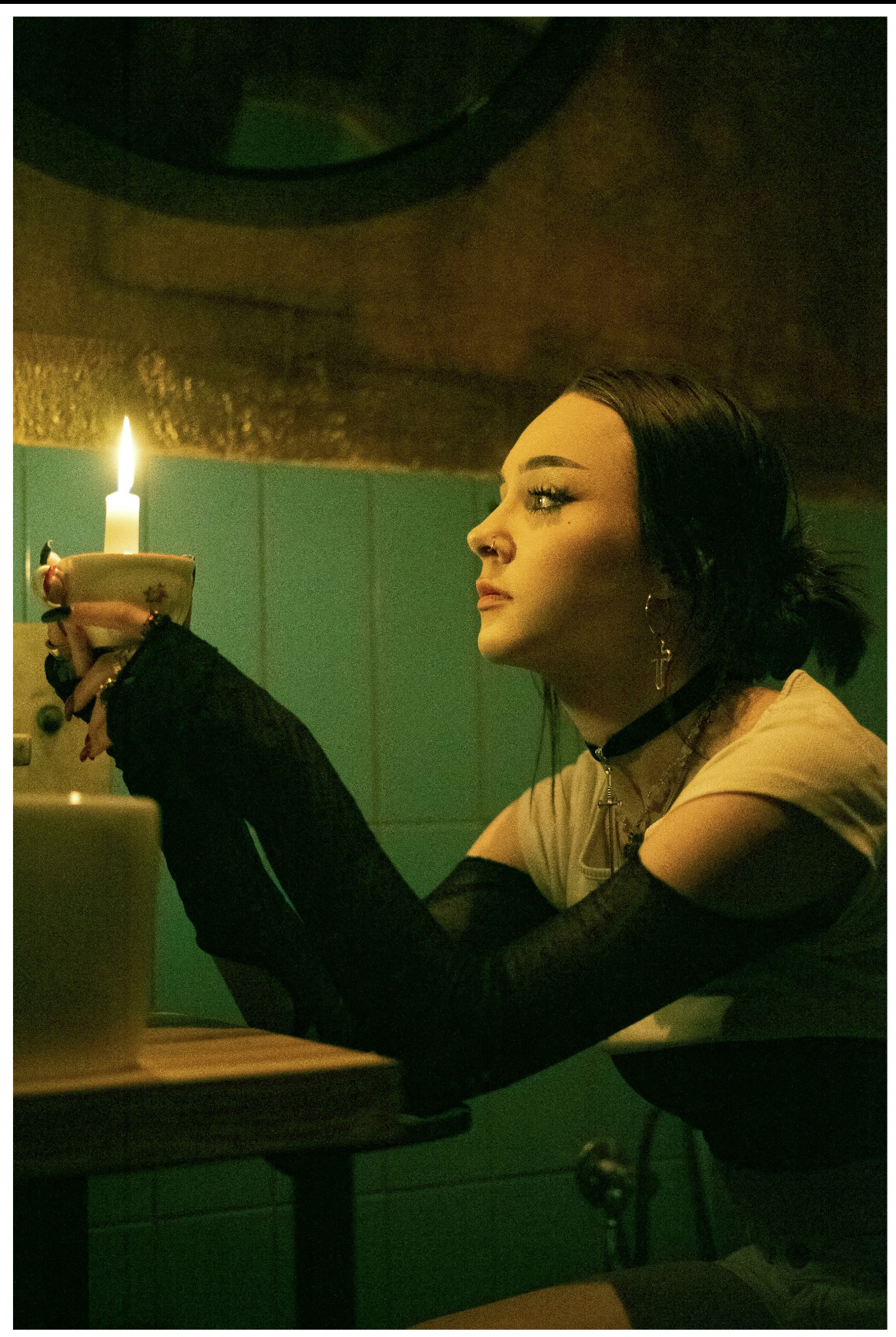 Portret kobiety w profilu, trzymającej świecę w ceramicznym kubku, z czarnymi rękawiczkami z koronki i chokerem z krzyżykiem, w retro oświetleniu i wnętrzu.