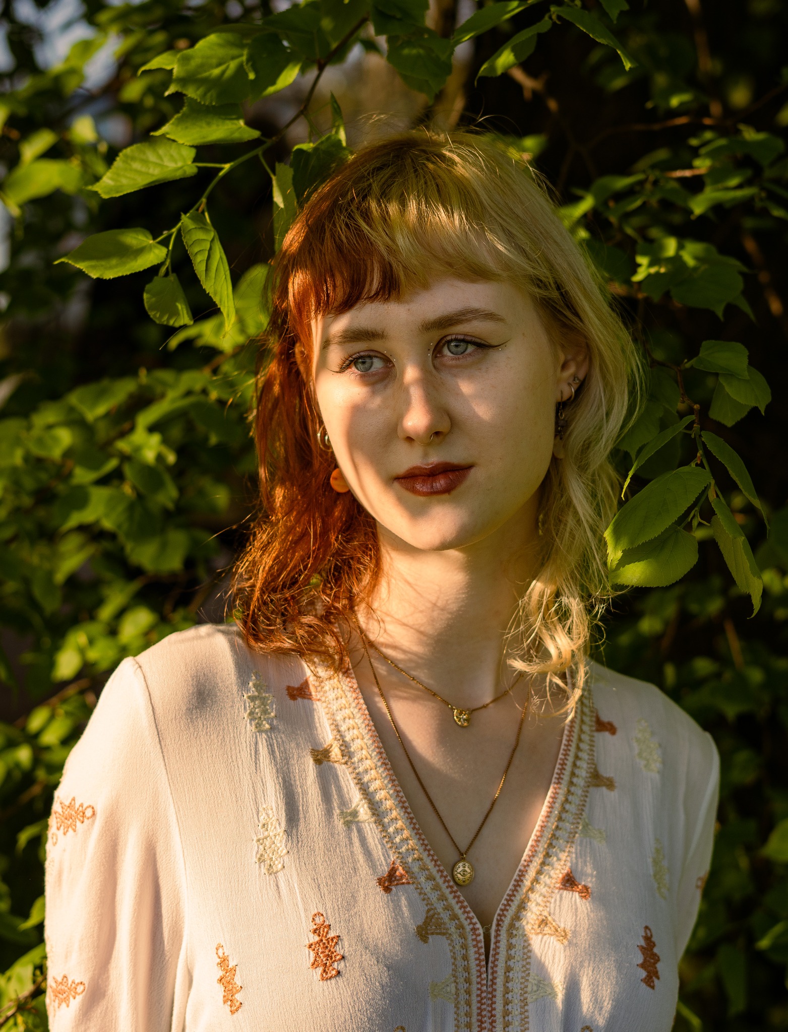 Portret młodej kobiety z dwukolorowymi włosami (blond i ruda) w naturalnym świetle, ubrana w haftowaną bluzkę i złotą biżuterię, pozująca na tle zielonych liści.
