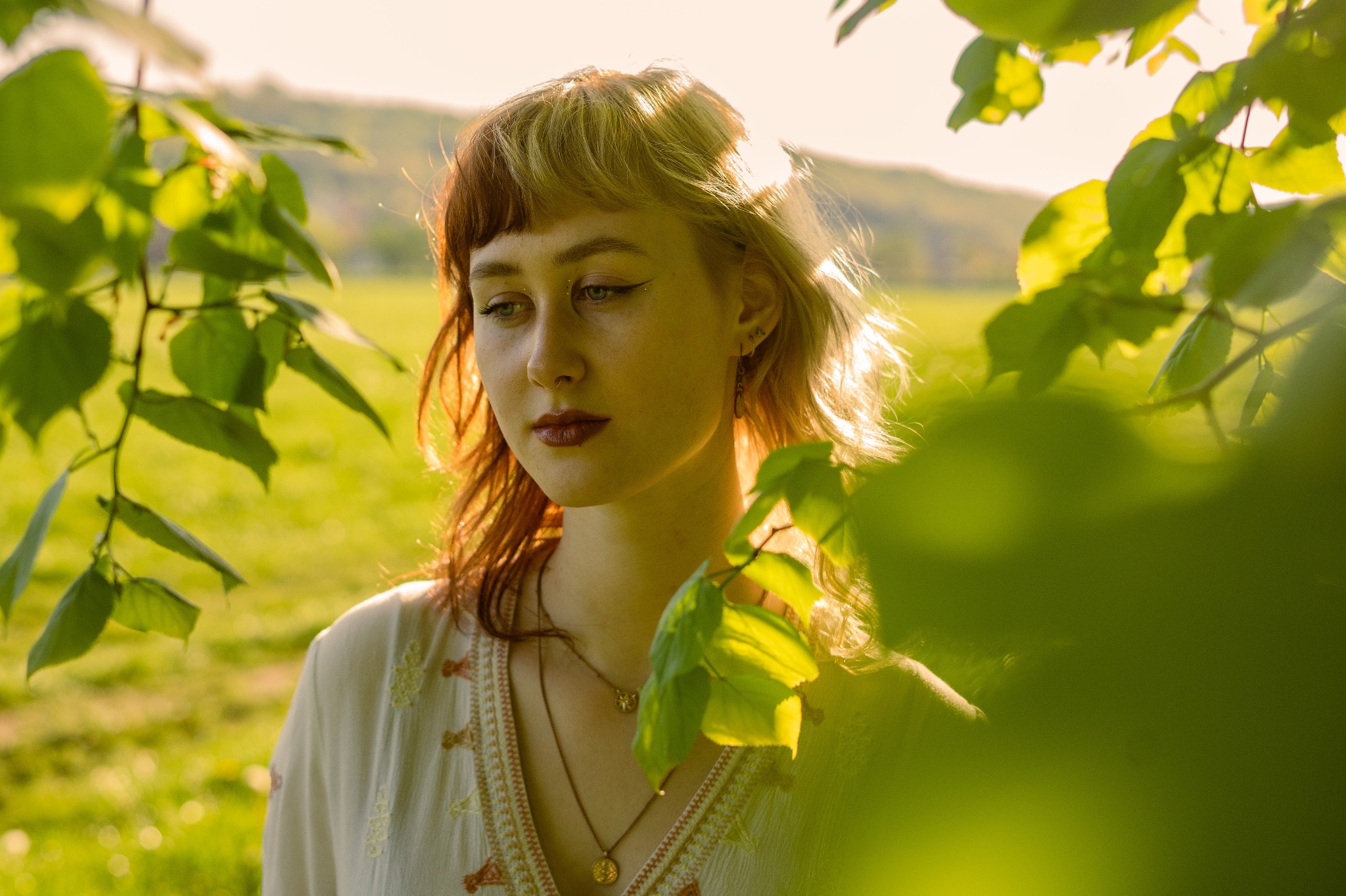 Portret kobiety z grzywką w stylu lat 70. w naturalnym świetle, z zielonymi liśćmi na pierwszym planie i łąką w tle.