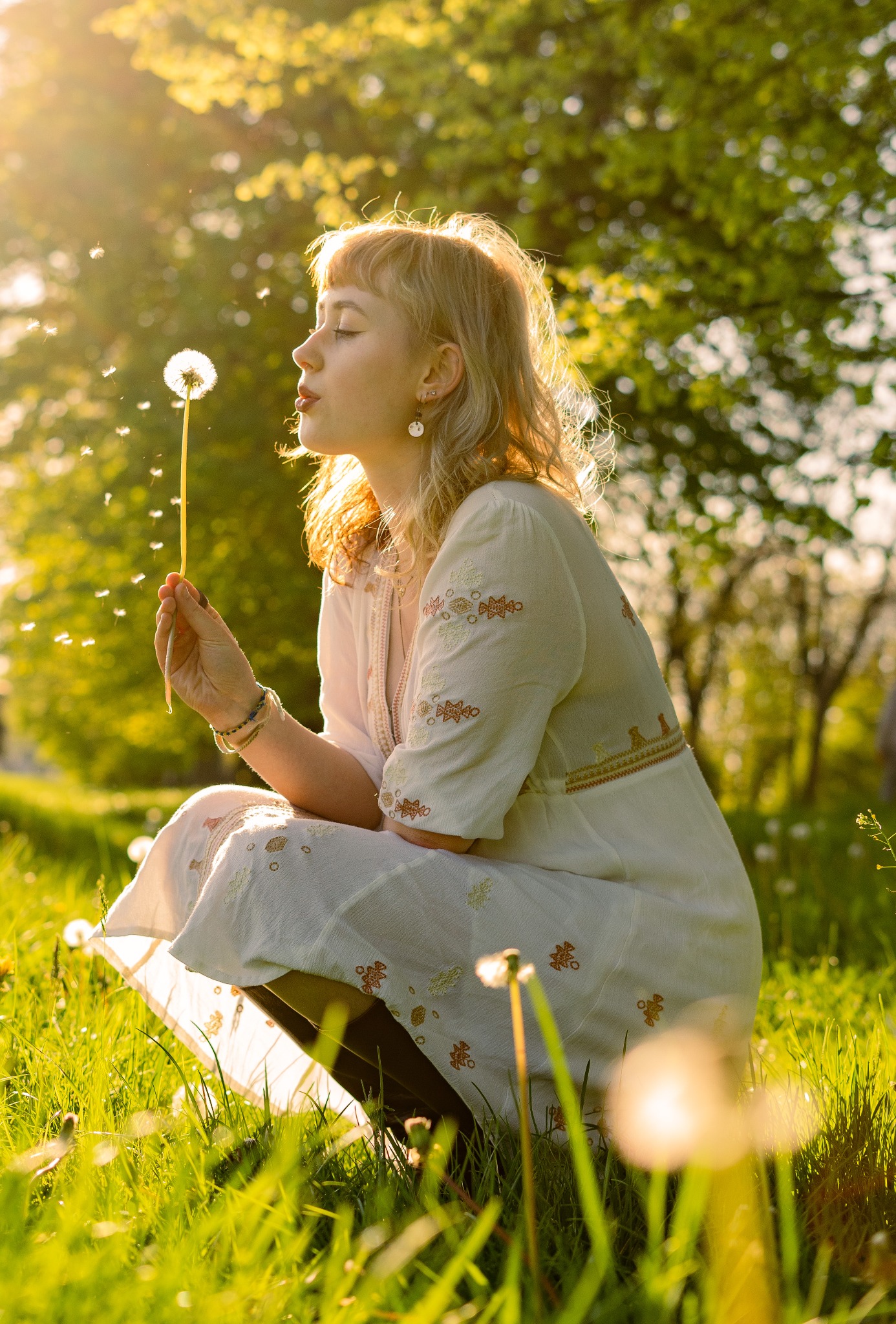 Kobieta w białej sukience dmucha na dmuchawca w słonecznym świetle, siedząc na łące; widoczne kolczyki i bransoletki.