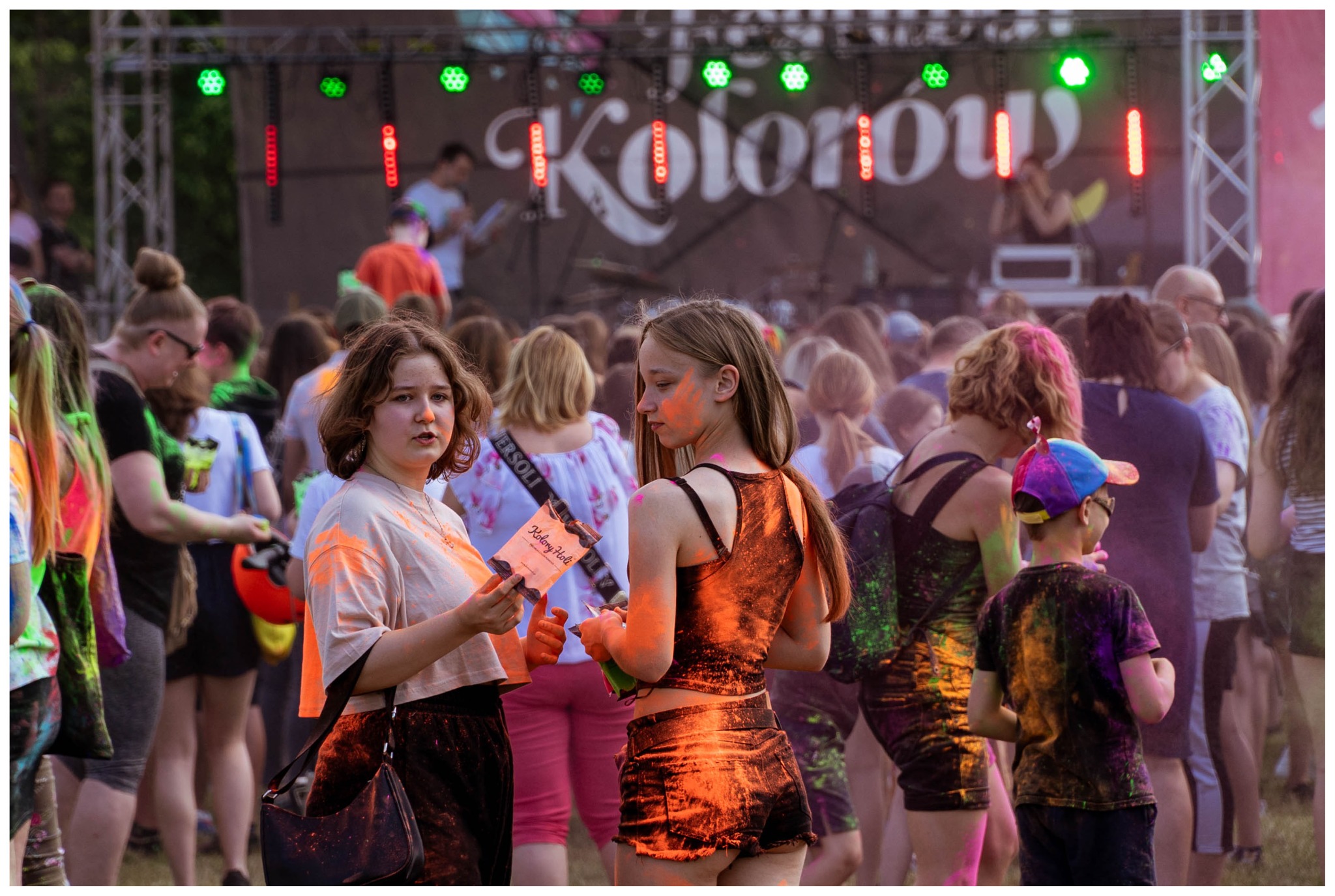 Tłum ludzi na festiwalu kolorów Holi, scena oświetlona zielonymi i czerwonymi światłami w tle, dziewczyna trzymająca ulotkę z napisem 'Kolory Holi'.