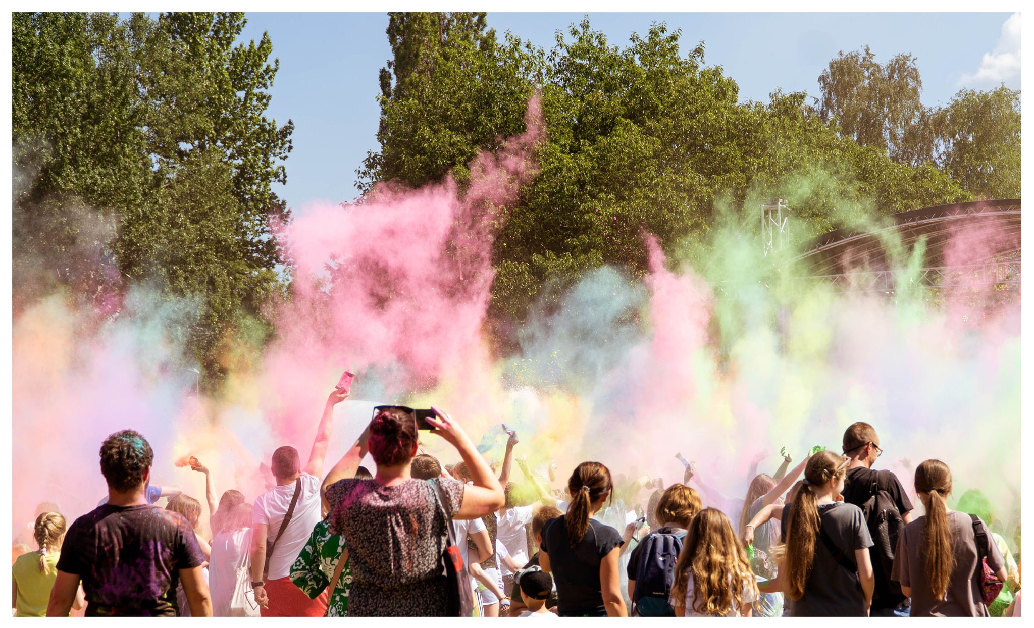Tłum ludzi podczas festiwalu kolorów Holi, unoszące się w powietrzu różowe i zielone proszki, drzewa w tle.