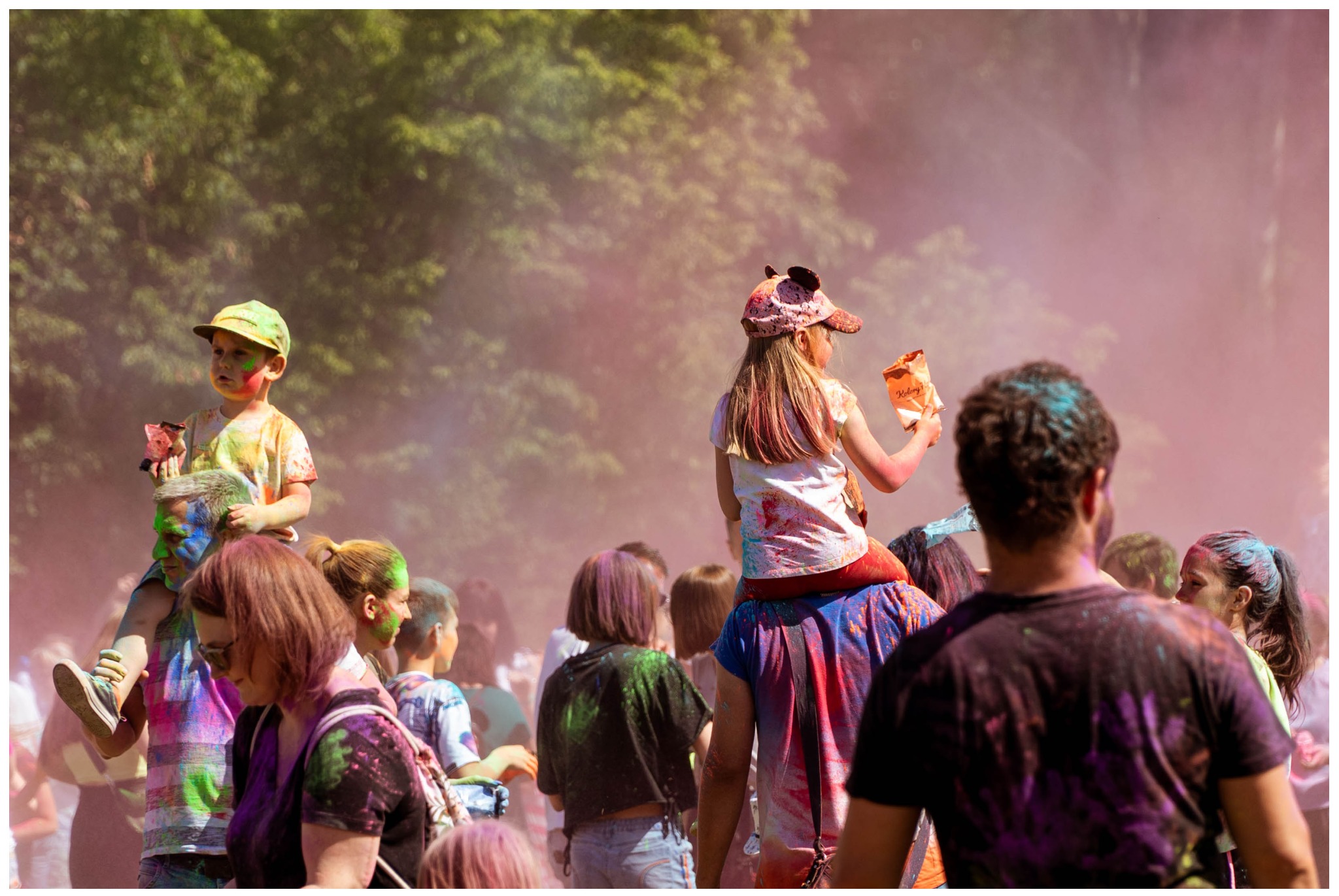 Tłum ludzi uczestniczących w festiwalu kolorów Holi, dzieci siedzą na ramionach rodziców, wszyscy pokryci kolorowym proszkiem, w tle drzewa i unoszący się pył.