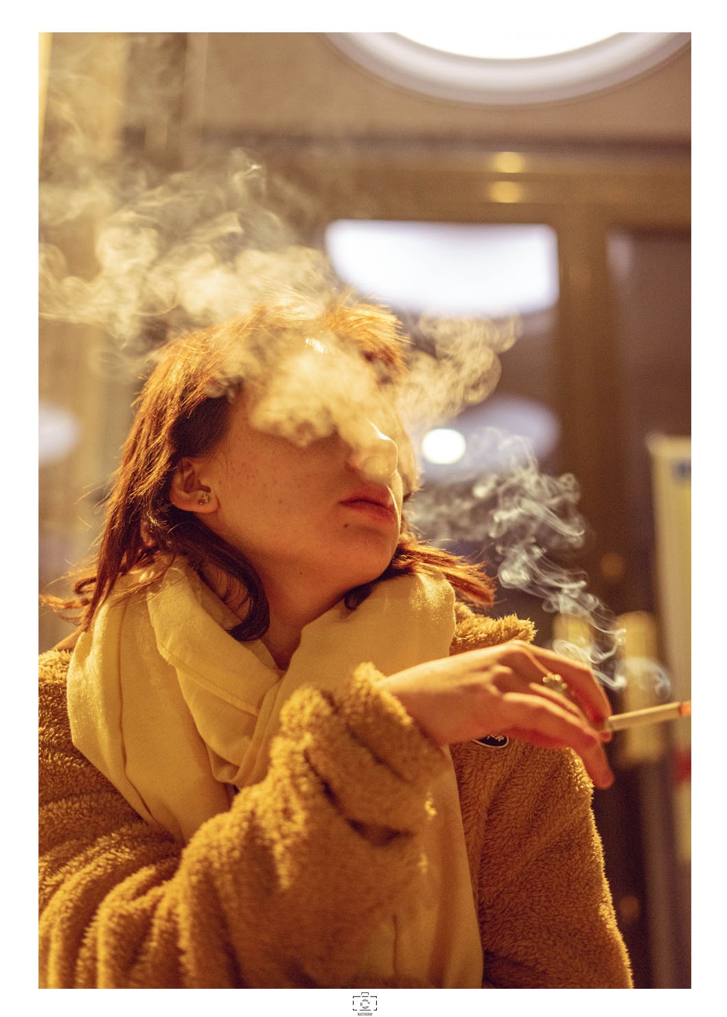 Portret kobiety z rudymi włosami, palącej papierosa, z dymem zasłaniającym twarz, ubrana w żółty szal i brązową kurtkę z futerka, w tle niewyraźne światła.