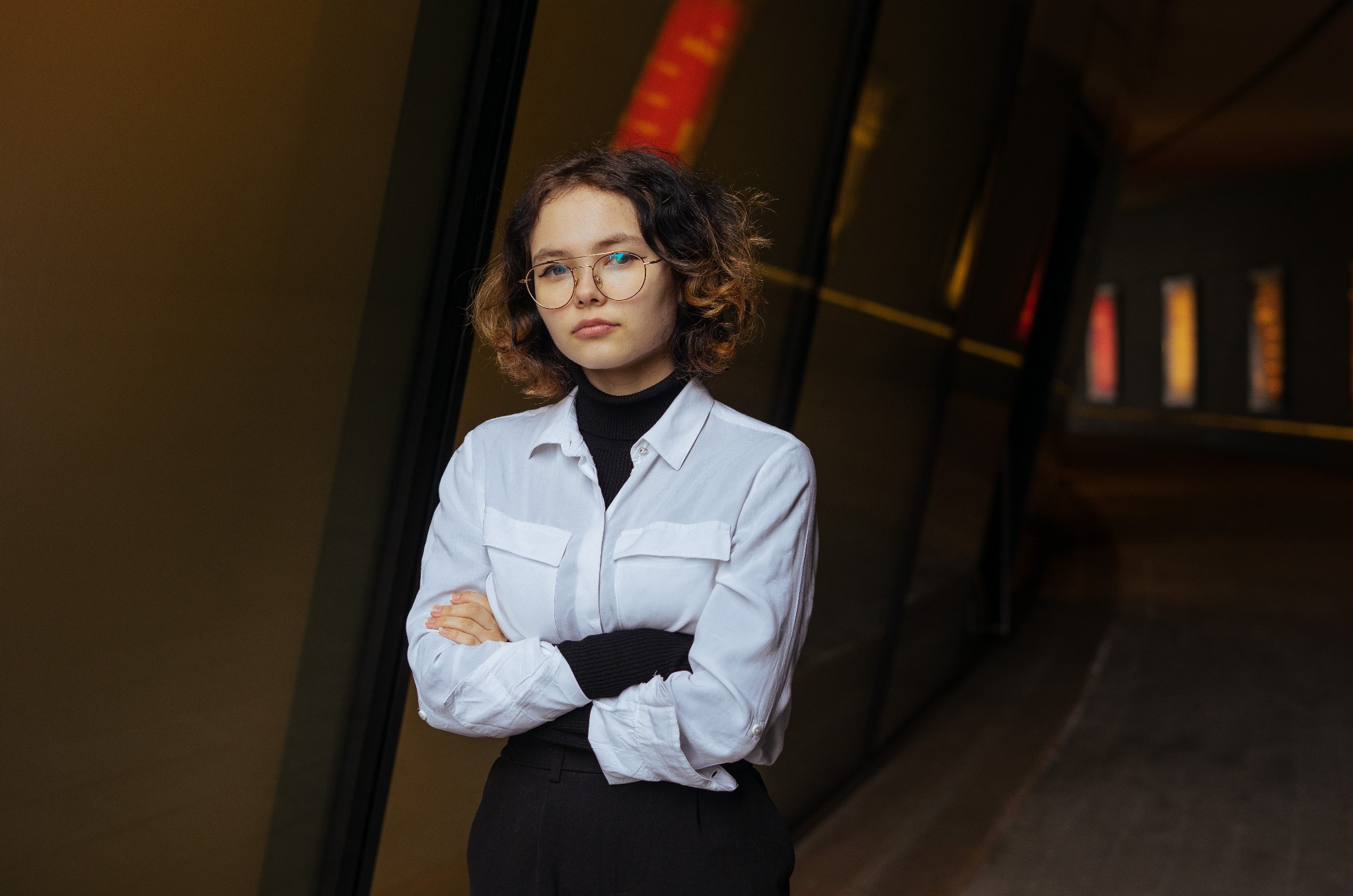 Portret młodej kobiety w okularach, ubrana w białą koszulę i czarny golf, pozująca na tle nowoczesnego budynku z ciemnymi panelami.