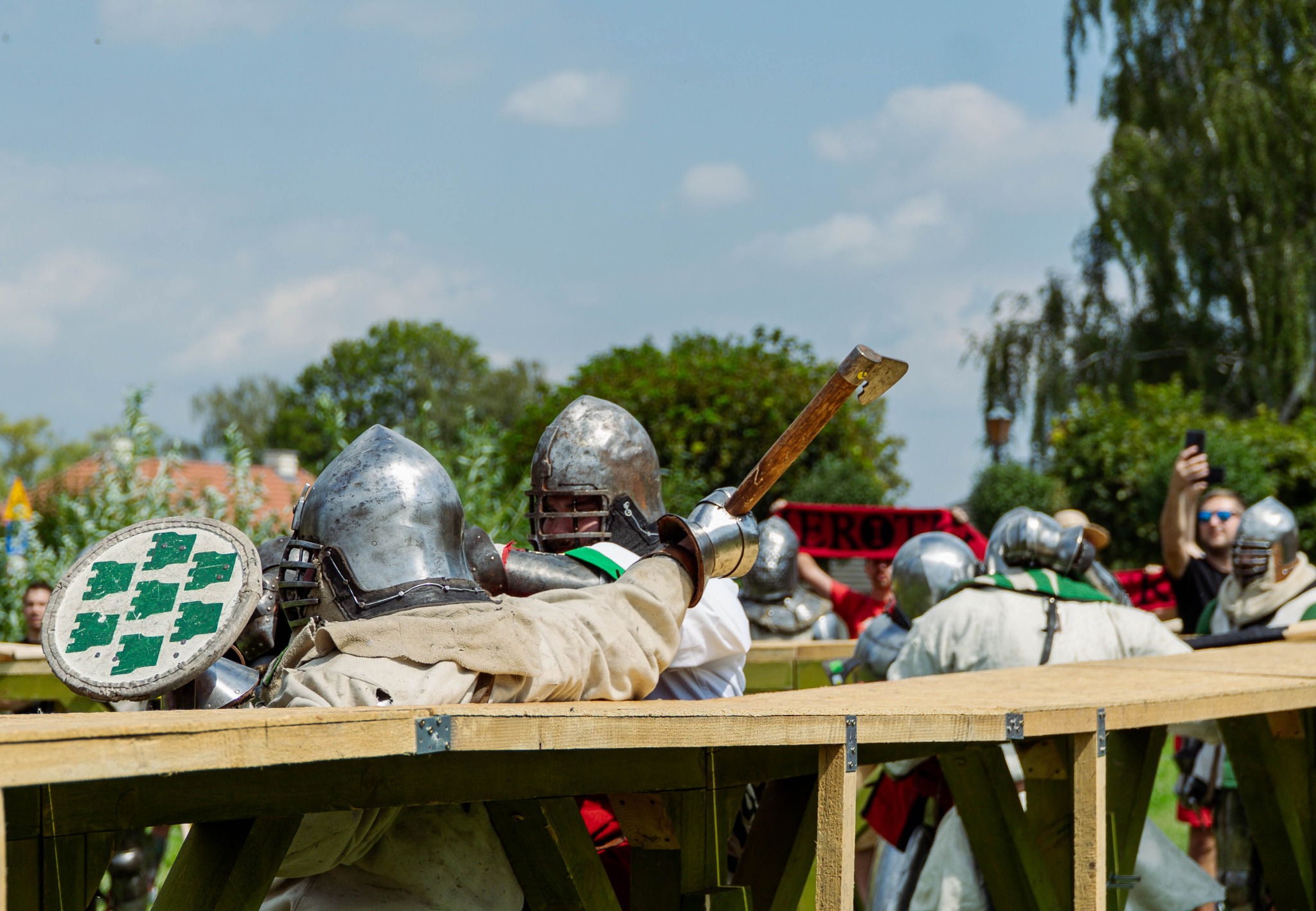 Grupa rekonstruktorów historycznych w pełnym uzbrojeniu średniowiecznym, walczących na drewnianej konstrukcji, z widocznym toporem w dłoni jednego z nich, fotografowanych przez osobę w tle.