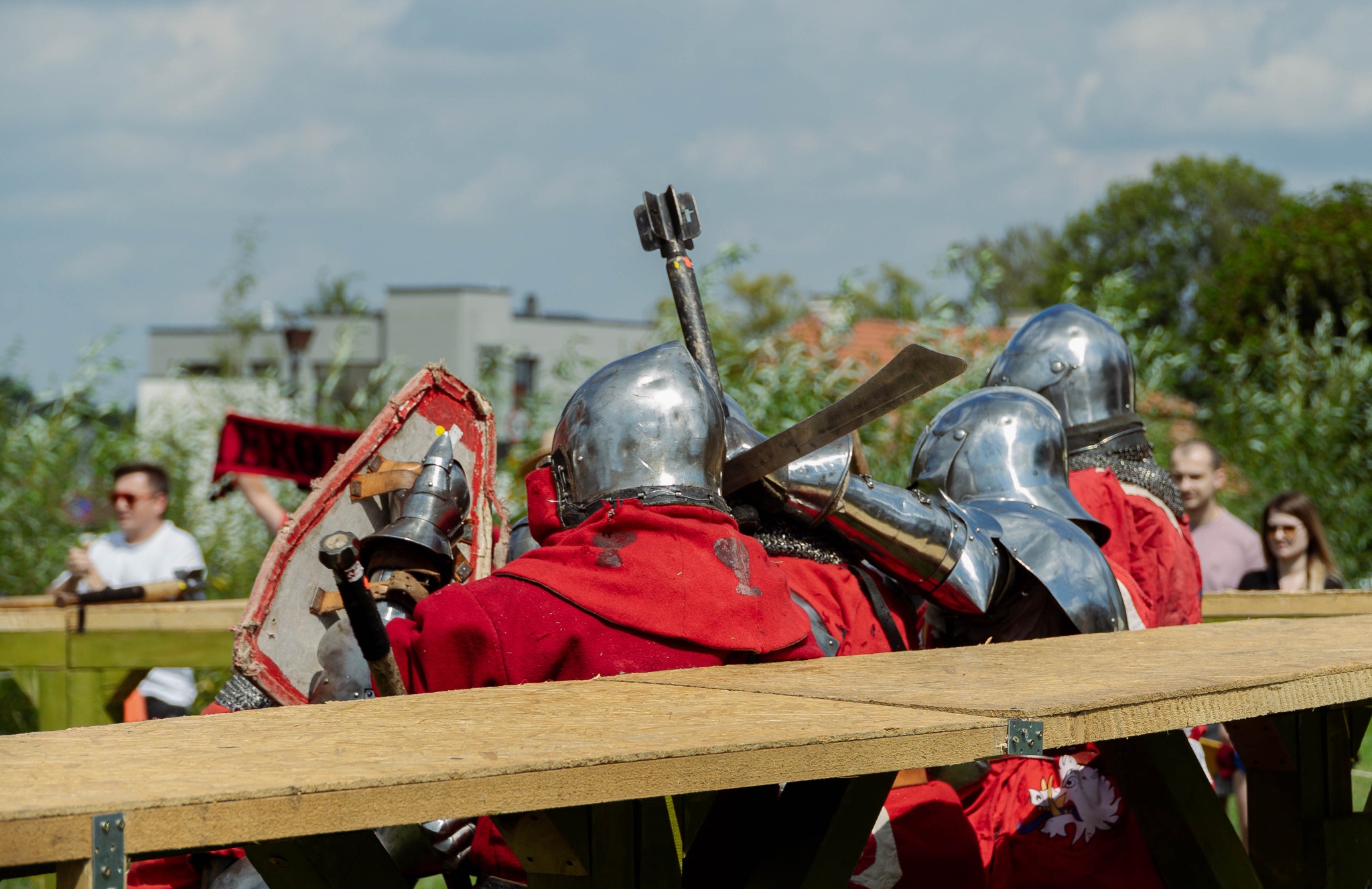 Grupa rycerzy w pełnym rynsztunku, z hełmami i bronią, stoi za drewnianą barierką, obserwując scenę w tle z publicznością.