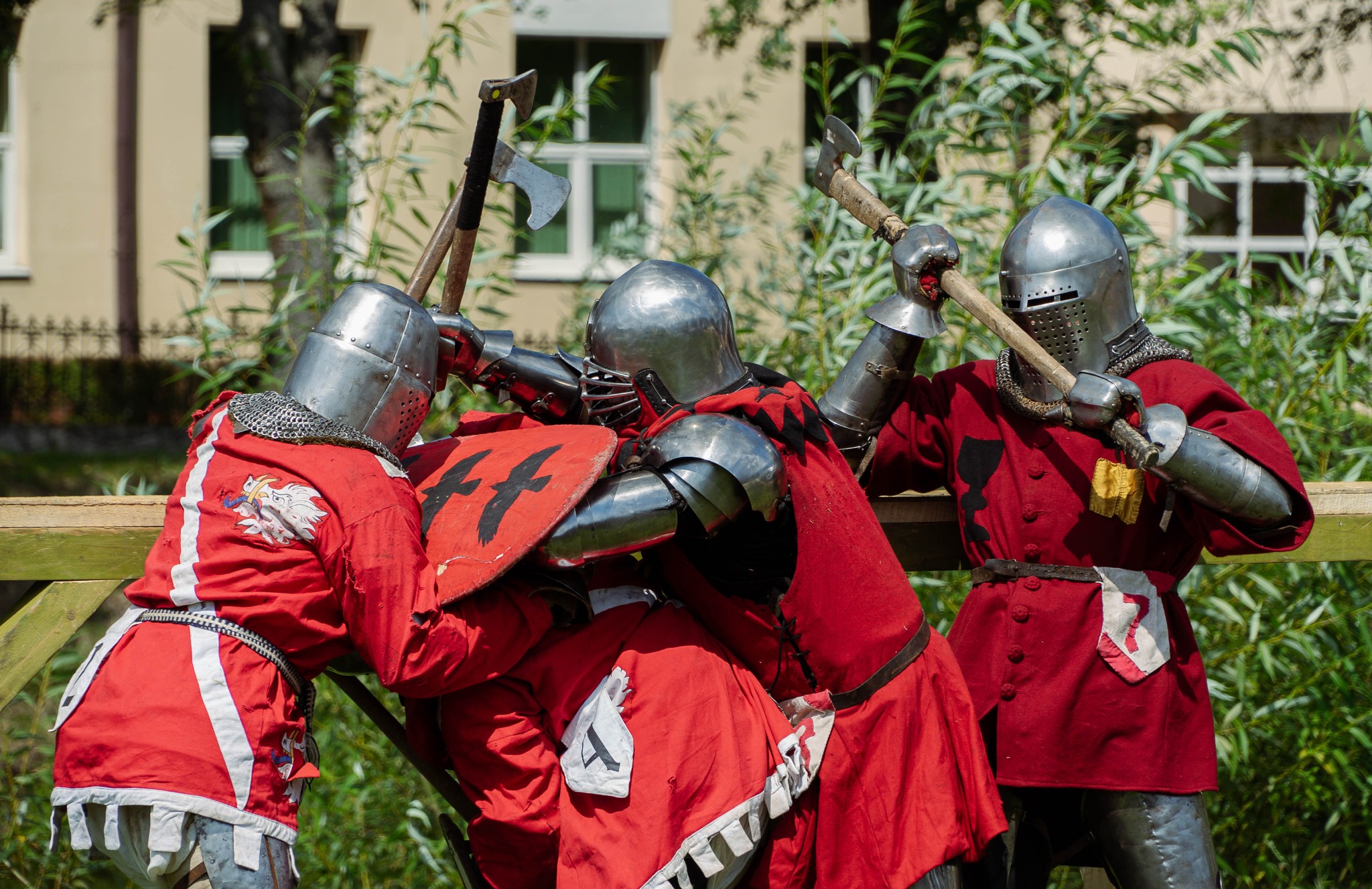 Rekonstrukcja historyczna: trzech rycerzy w pełnym uzbrojeniu, z toporami i tarczą, w trakcie walki na tle budynku.