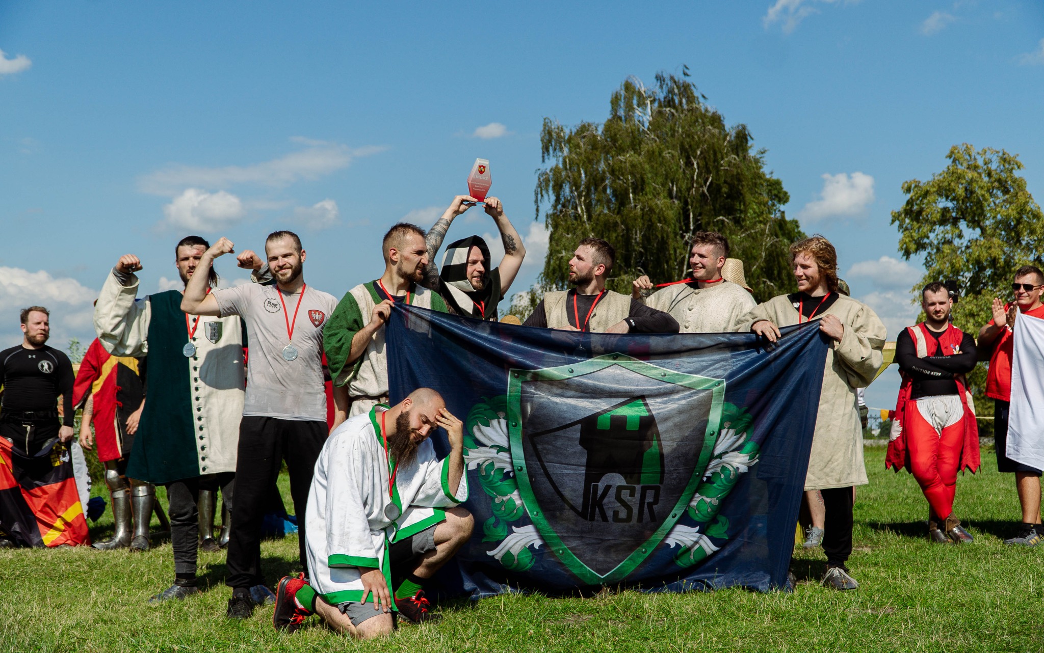Grupa mężczyzn w strojach stylizowanych na historyczne, trzymających flagę z logiem KSR i świętujących zwycięstwo, jeden z nich trzyma trofeum.