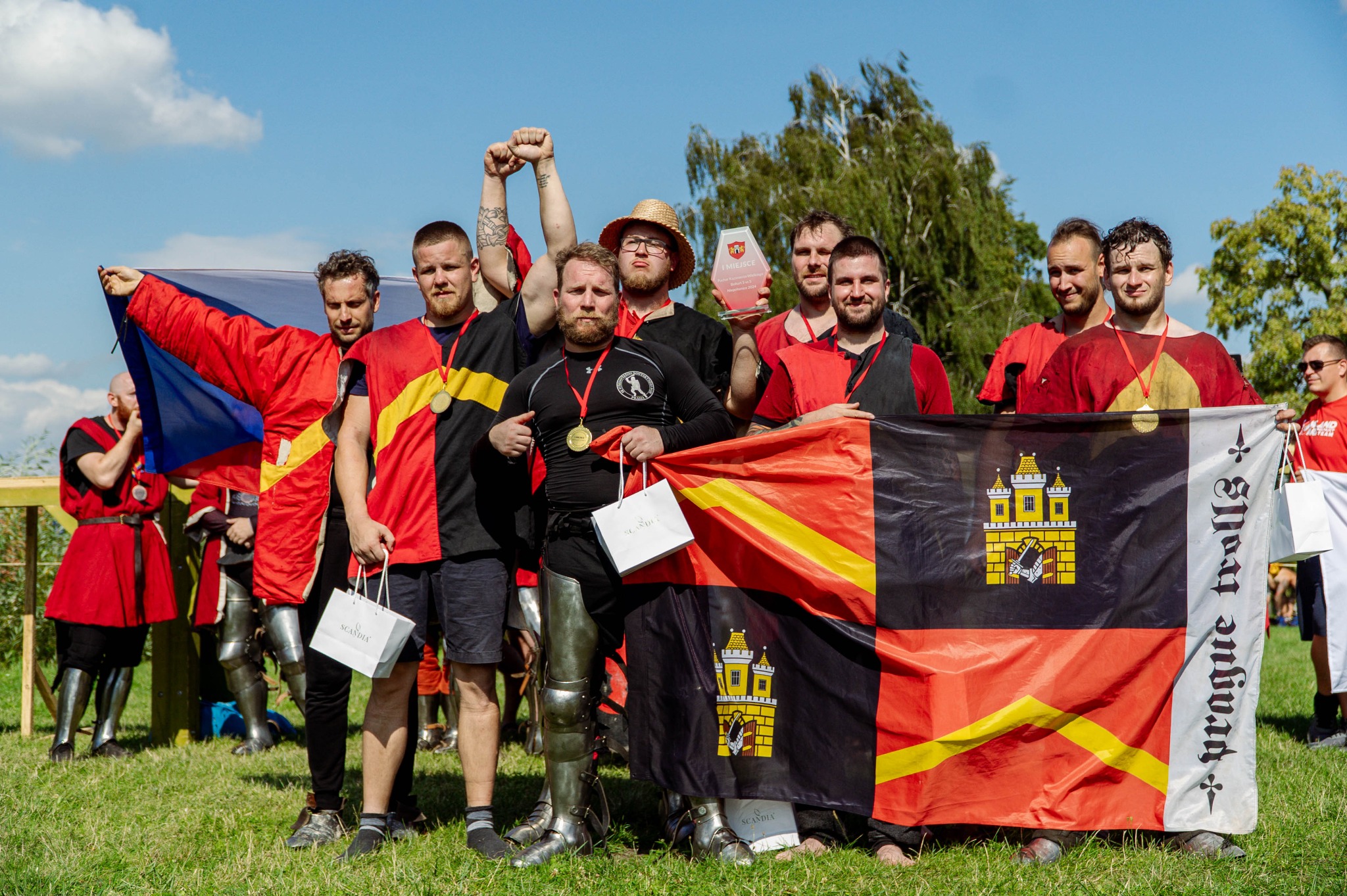 Drużyna rycerska 'Prague trolls' świętuje zwycięstwo na trawie, trzymając flagę z herbem i trofeum, w strojach turniejowych z medalami.