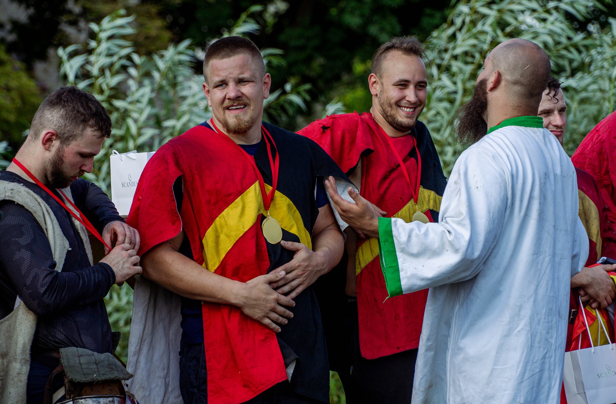 Grupa mężczyzn w kostiumach z epoki, z medalami na szyi, rozmawia na tle zieleni.