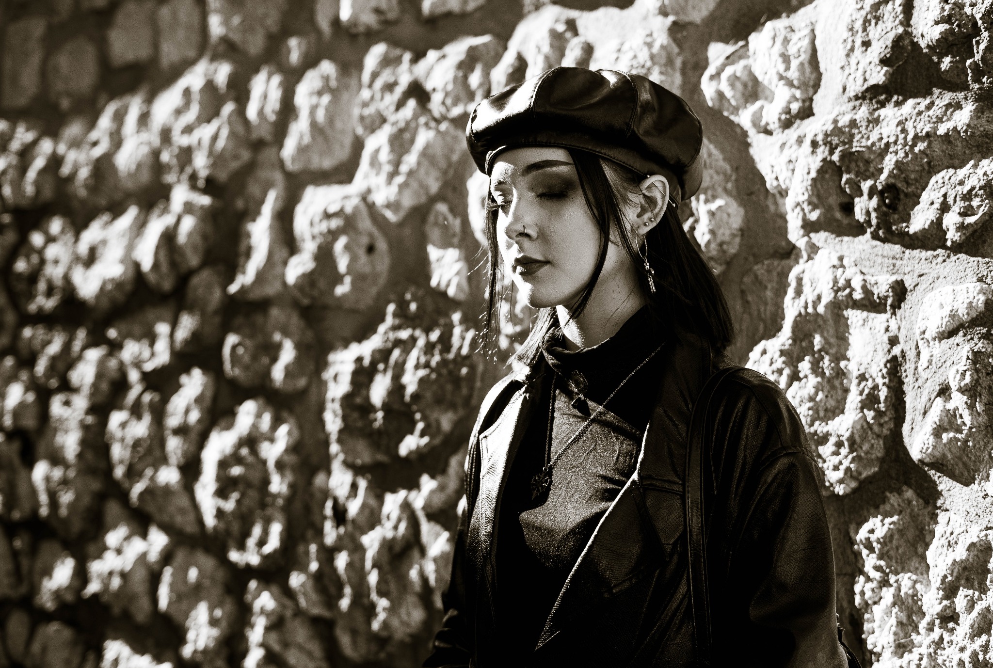 Portret kobiety w czarnej skórzanej kurtce i beretce, opierającej się o kamienną ścianę w sepii, z zamkniętymi oczami i delikatnym oświetleniem bocznym podkreślającym teksturę ściany.