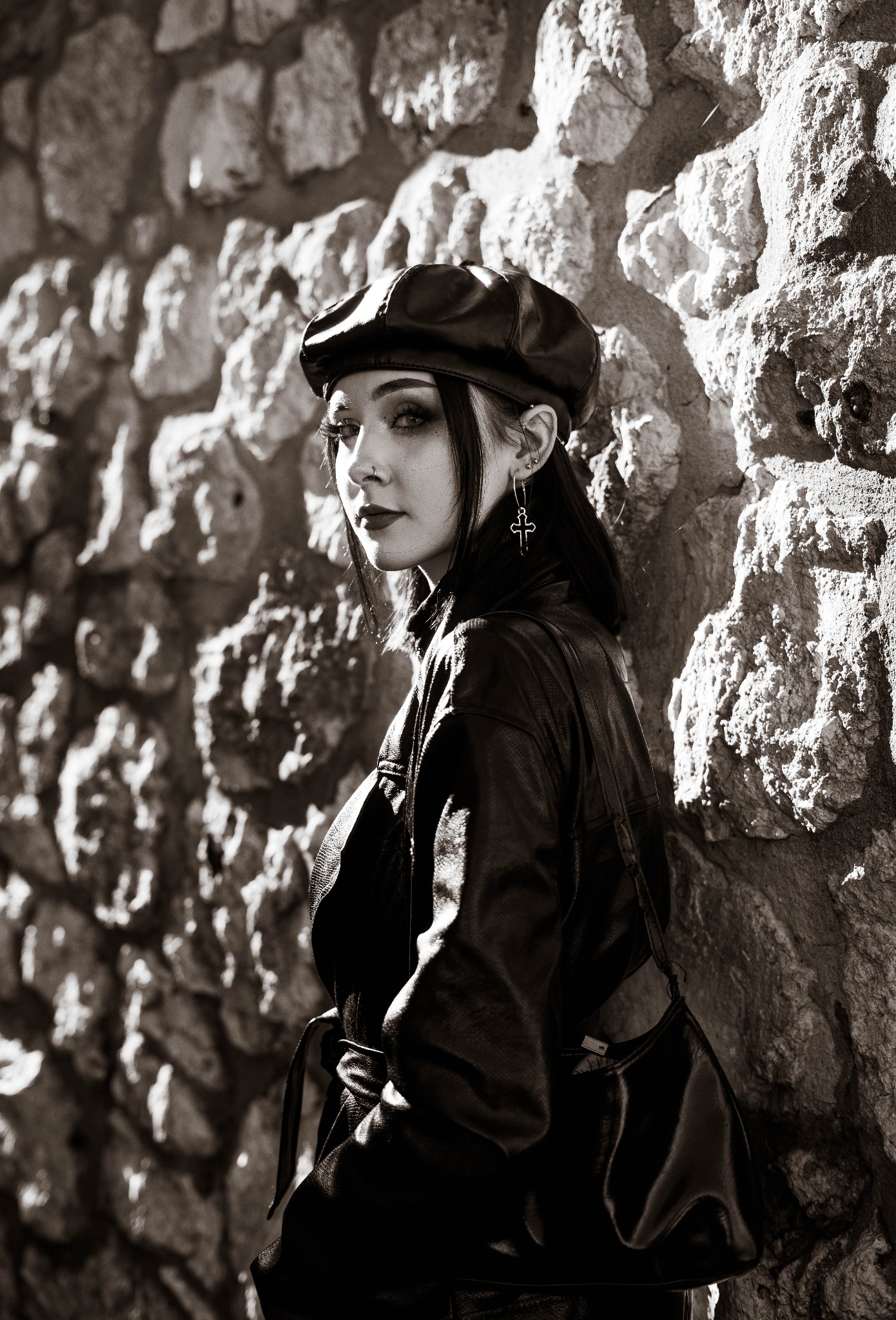 Czarno-białe zdjęcie kobiety w skórzanej kurtce i beretce, opierającej się o kamienną ścianę, z kolczykiem w kształcie krzyża.