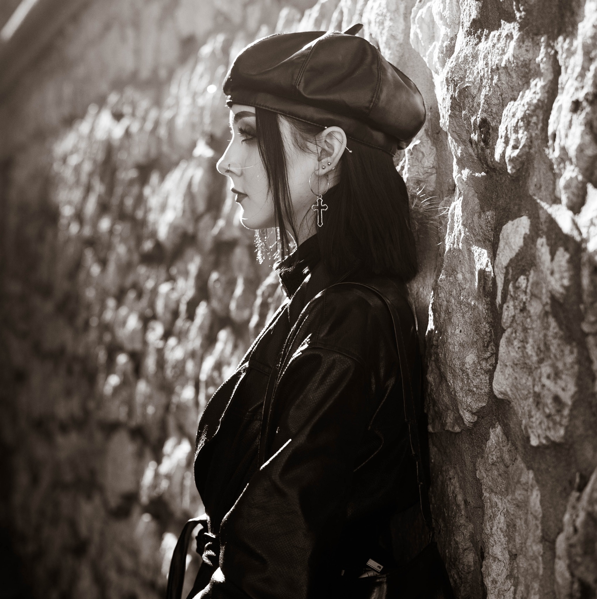 Czarno-białe zdjęcie profilu kobiety w skórzanym berecie, kurtce i kolczykach z krzyżami, opierającej się o kamienną ścianę w ostrym świetle.