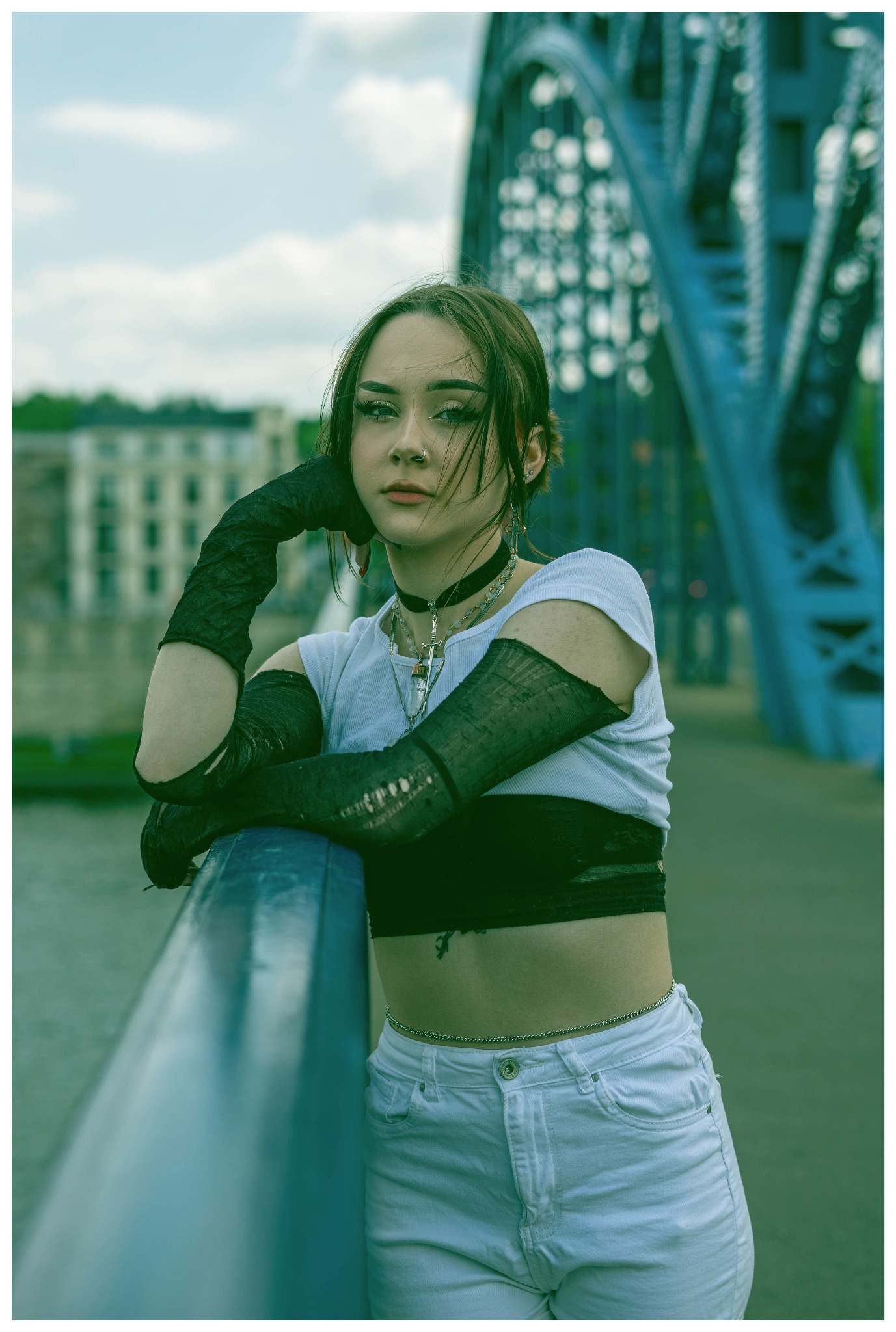 Portret młodej kobiety w gotyckim stylu, opierającej się o barierę mostu na tle metalowej konstrukcji i zabudowy miejskiej.